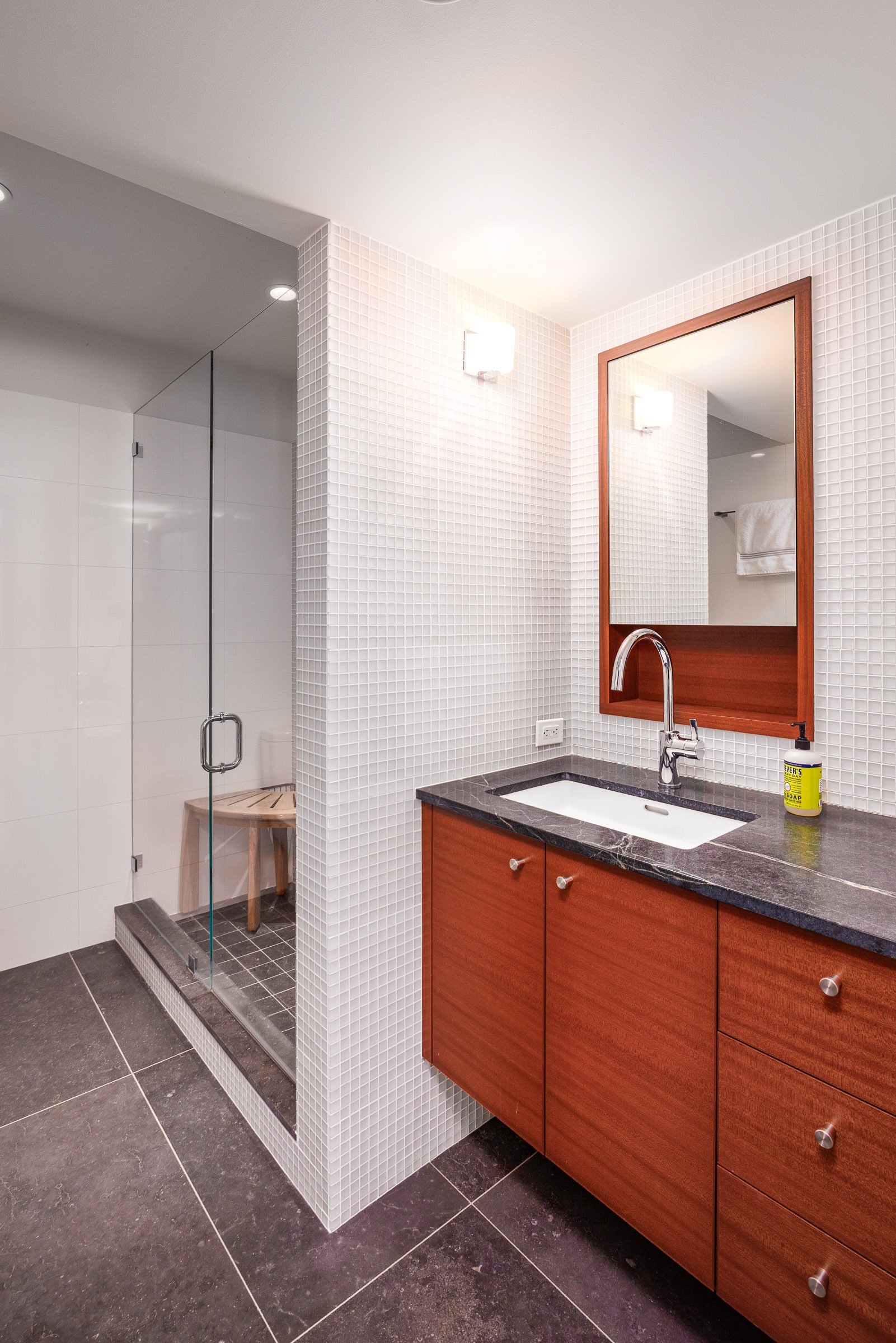 Modern bathroom with glass-enclosed shower, wooden vanity with black countertop, large mirror, wall-mounted lights, and a white tiled wall.