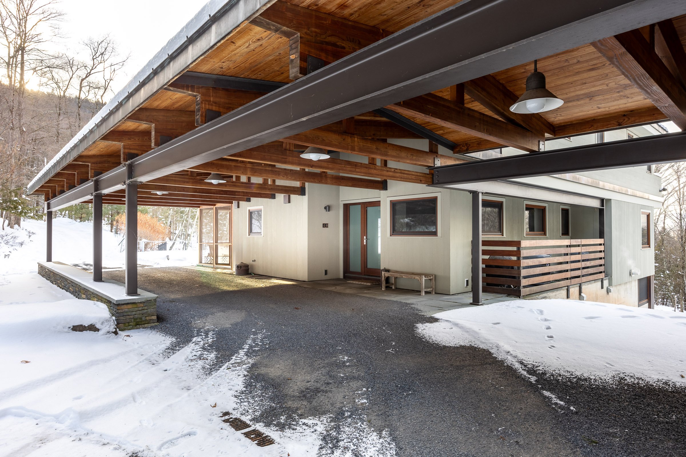 Modern house with a large wooden balcony overhang, supported by black metal beams, in a snowy landscape with bare trees.