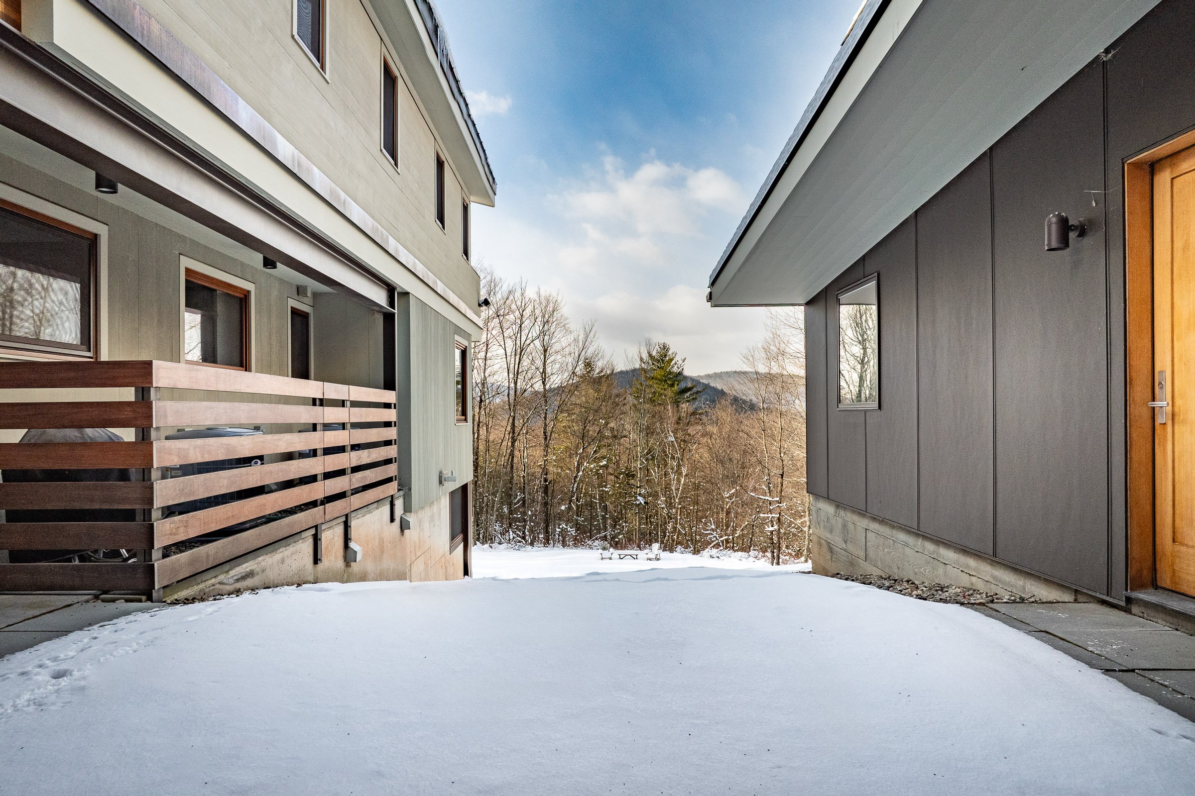 Snow-covered patio between two modern houses with large windows and wooden accents, surrounded by leafless trees and distant mountains under a blue sky with some clouds.