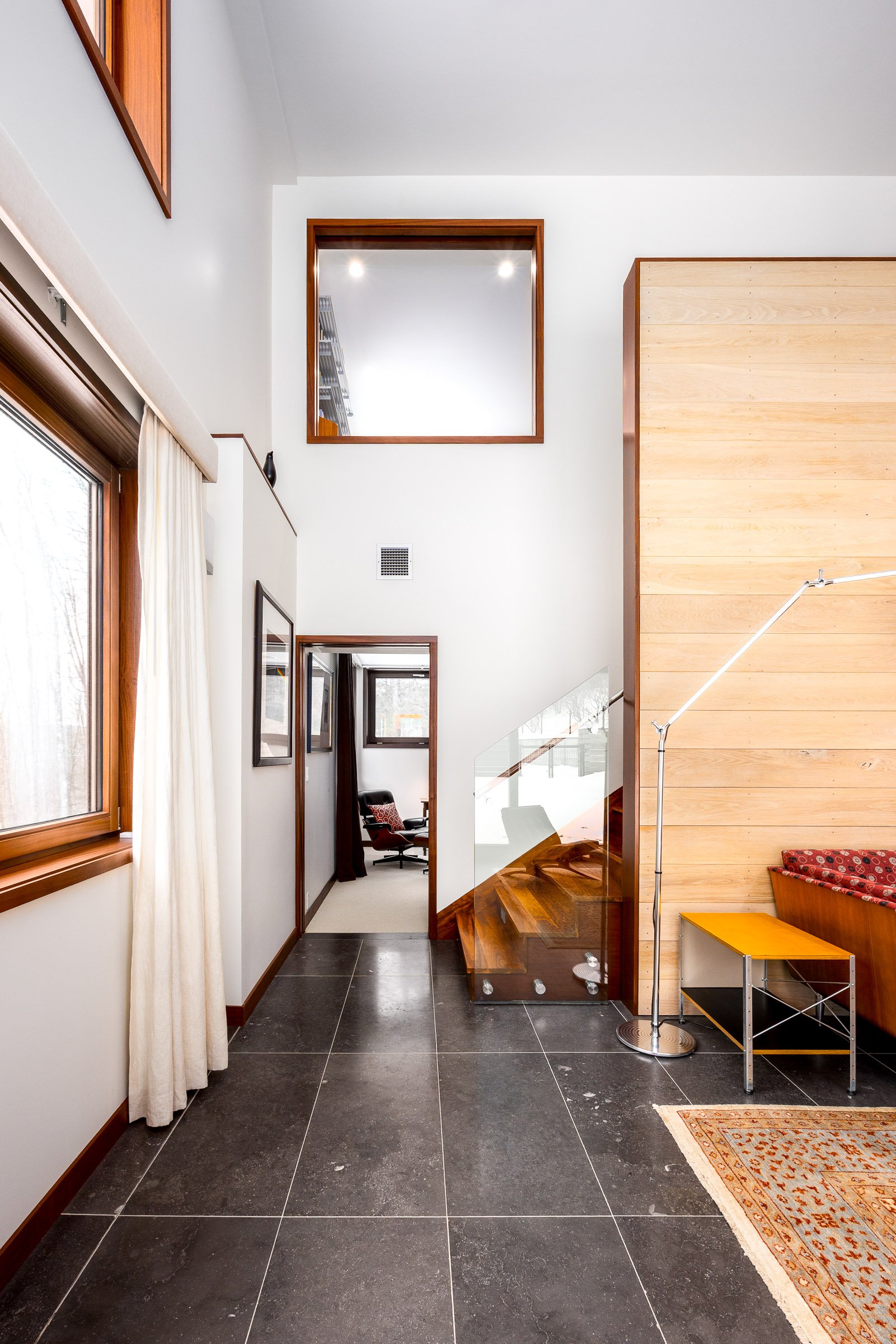 Modern interior hallway with large windows, a staircase with glass railing, and a sitting room with a purple chair visible through a doorway.