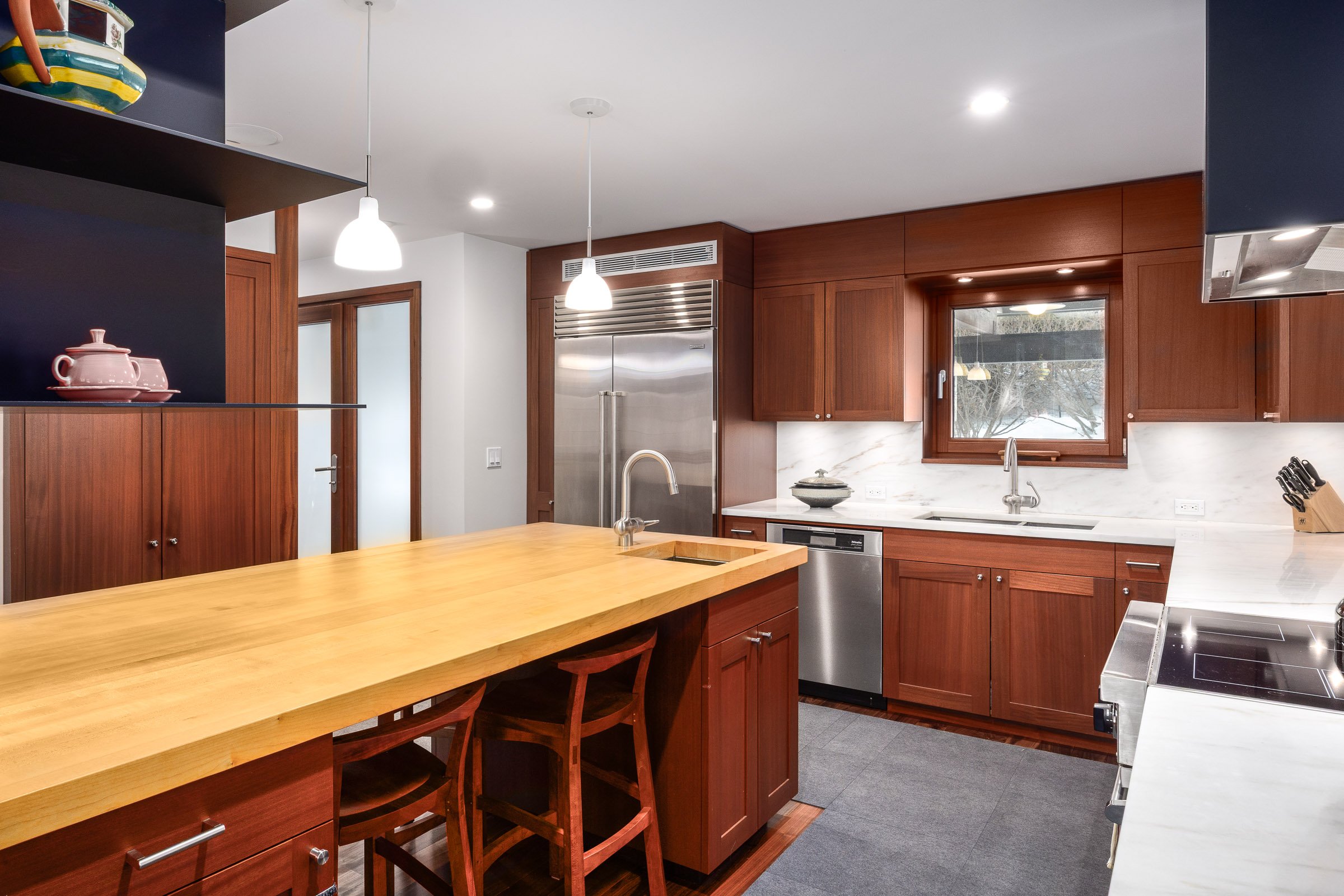 A modern kitchen with wooden cabinets, a marble backsplash, stainless steel appliances, a wooden kitchen island with two chairs, a window above the sink, and pendant lighting.