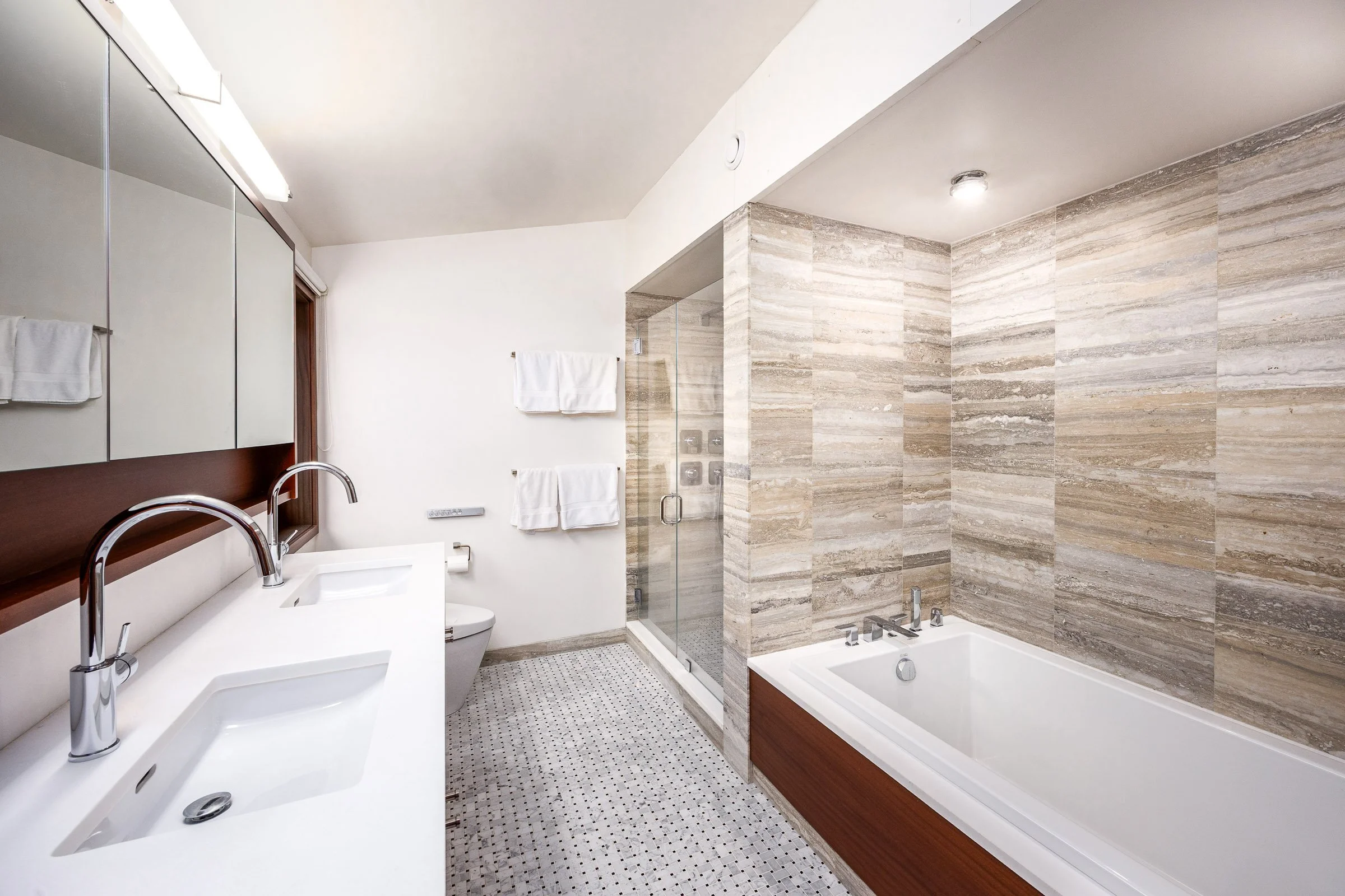 Modern bathroom with dual sinks, large mirror, toilet, glass shower enclosure, and bathtub with wood paneling, tiled walls, and patterned floor.
