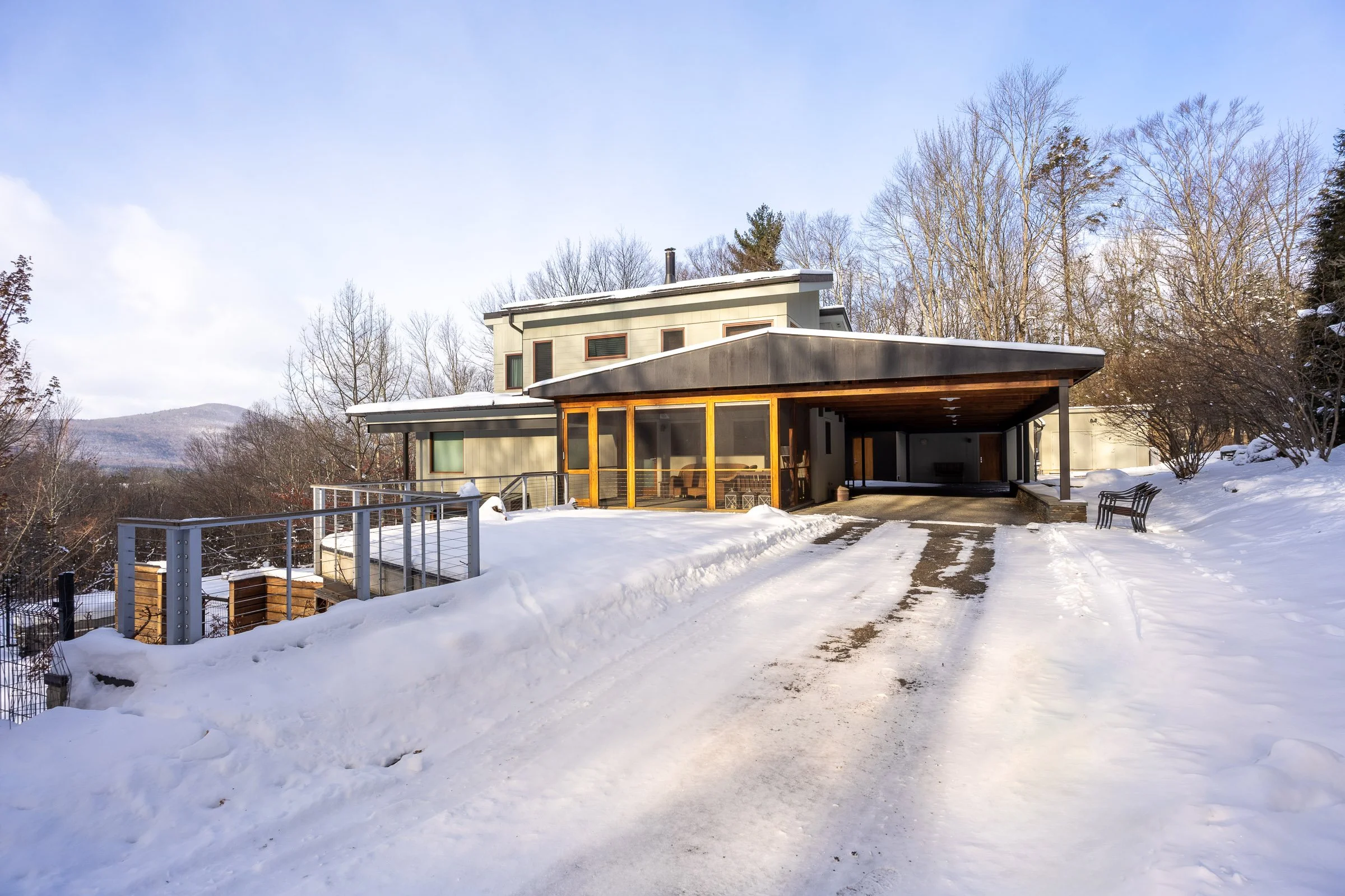 Modern house with large glass windows and a carport, surrounded by snow and leafless trees, in a winter landscape.