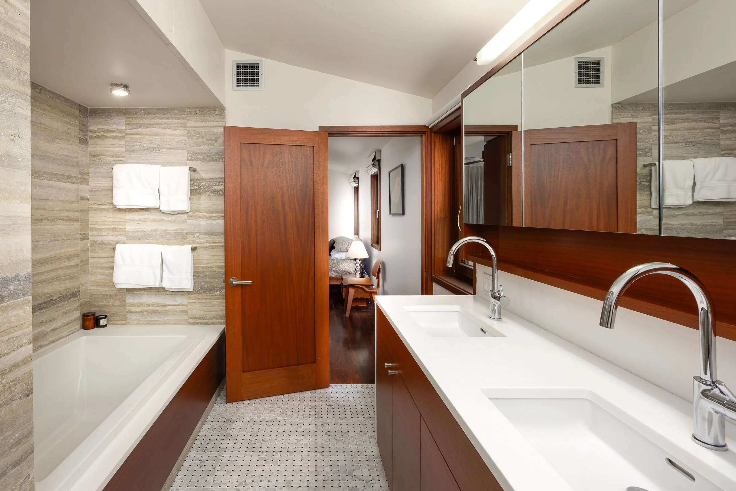 A modern bathroom with twin sinks, a large mirror, a bathtub, beige tiled walls, a wooden door open to a bedroom, and white towels hanging on the wall.