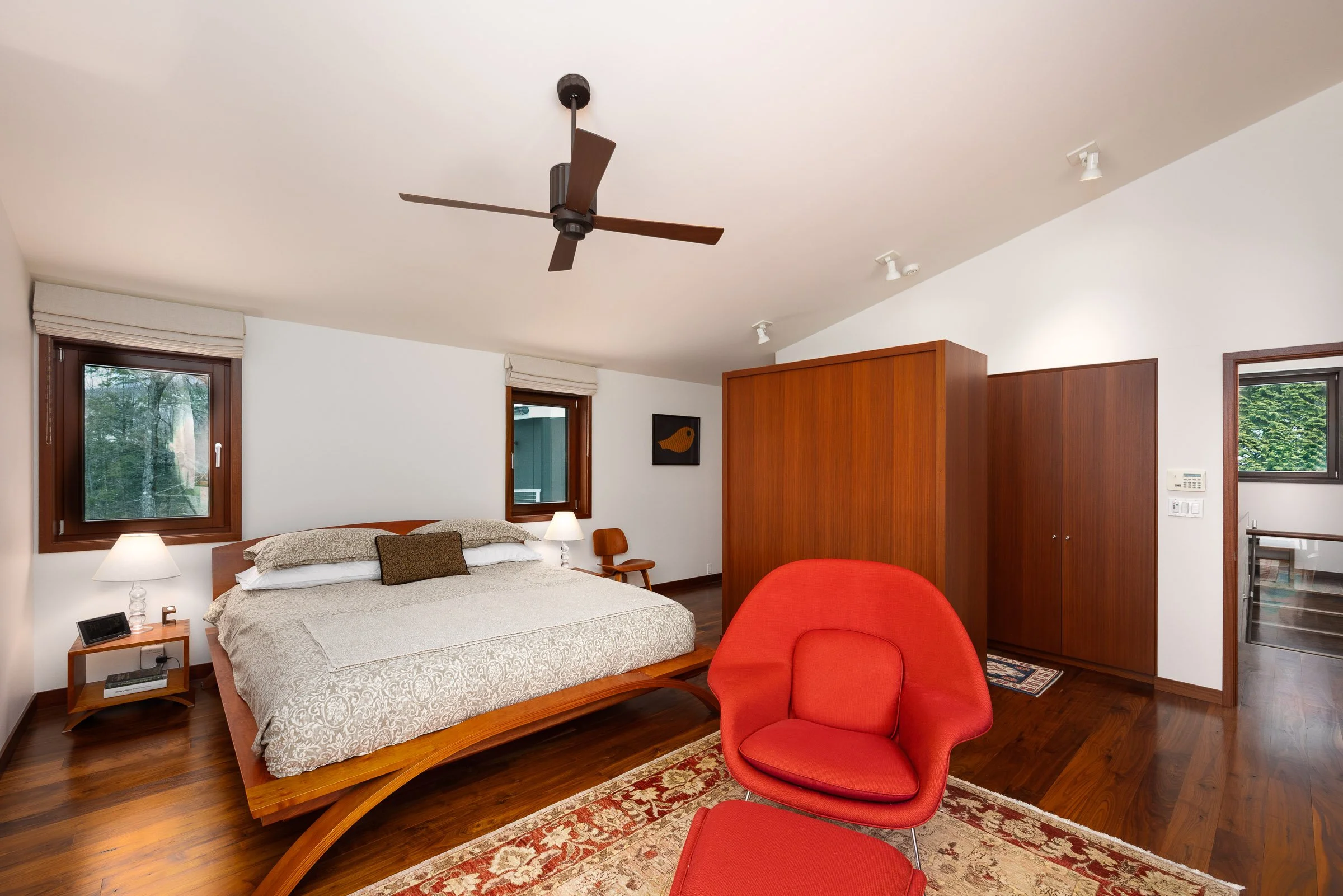 A bedroom with a large bed, side tables with lamps, two windows, a wooden wardrobe, a red armchair, and a ceiling fan.