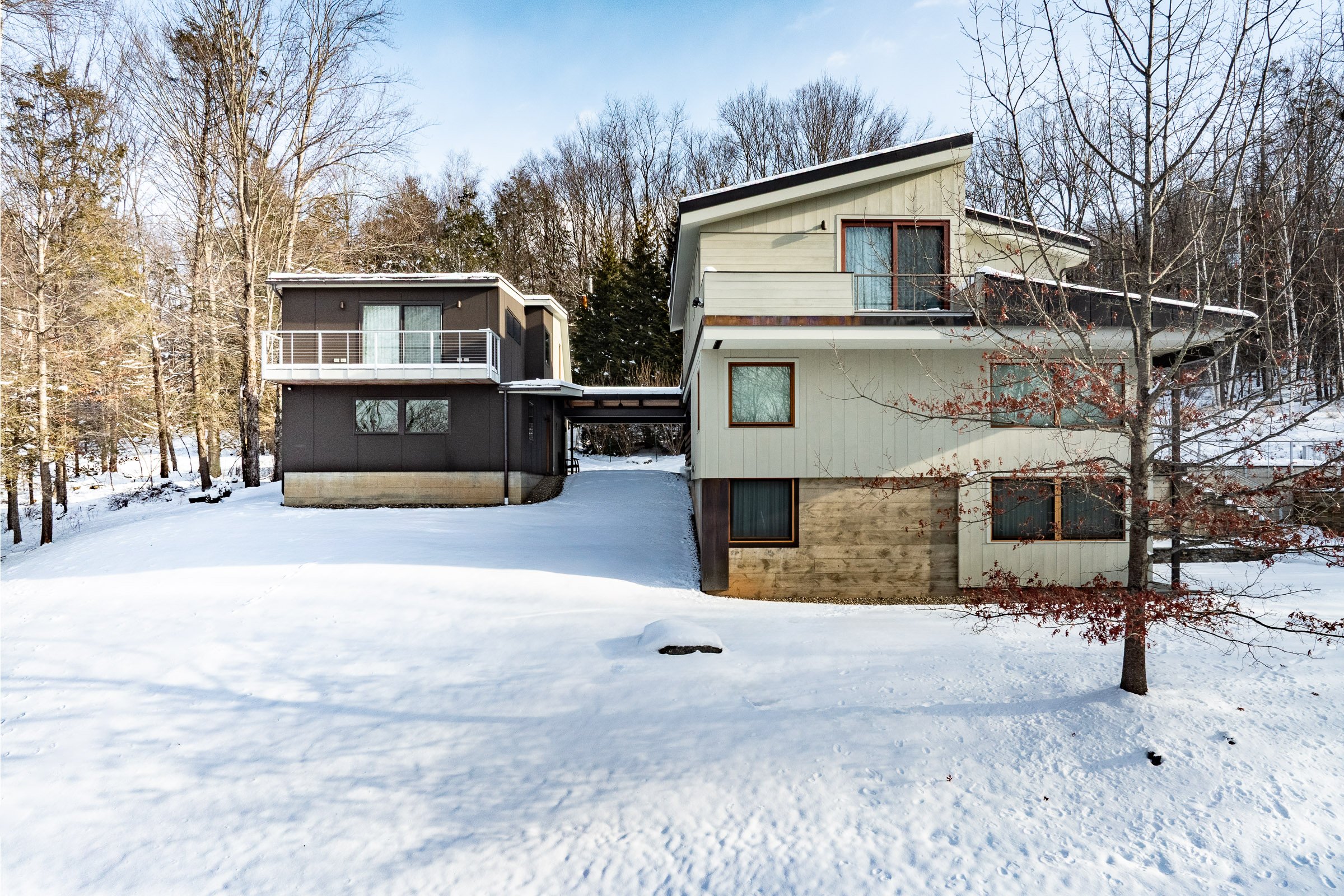 Modern house with multiple levels and large windows surrounded by snow and leafless trees in winter.