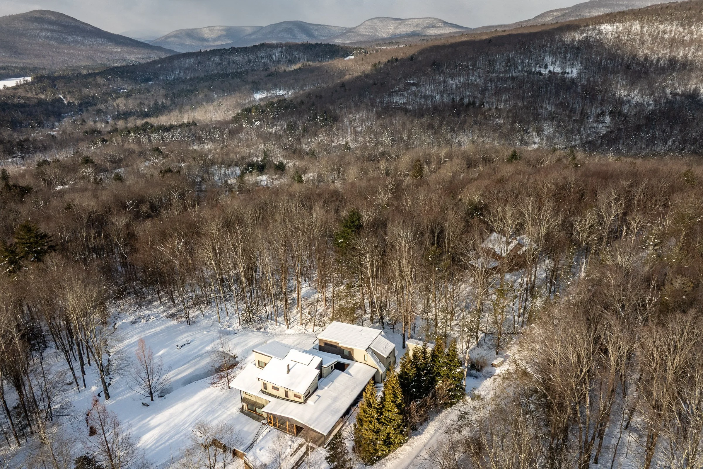 Bird's eye view of a house surrounded by snow-covered trees and mountains in the background during winter.
