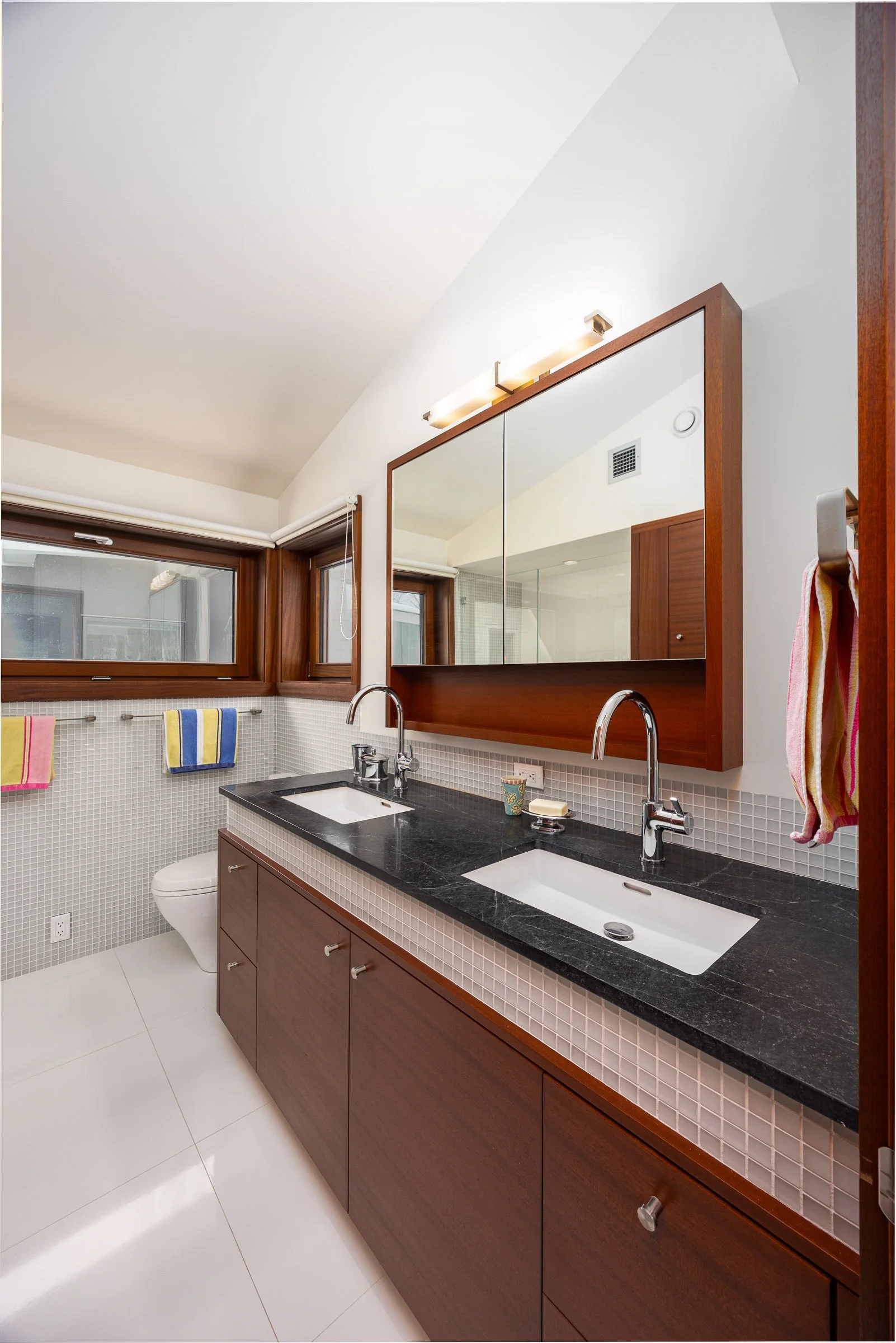 Modern bathroom with dual sinks, black countertop, large mirror, wood cabinets, window, towels, and white fixtures.