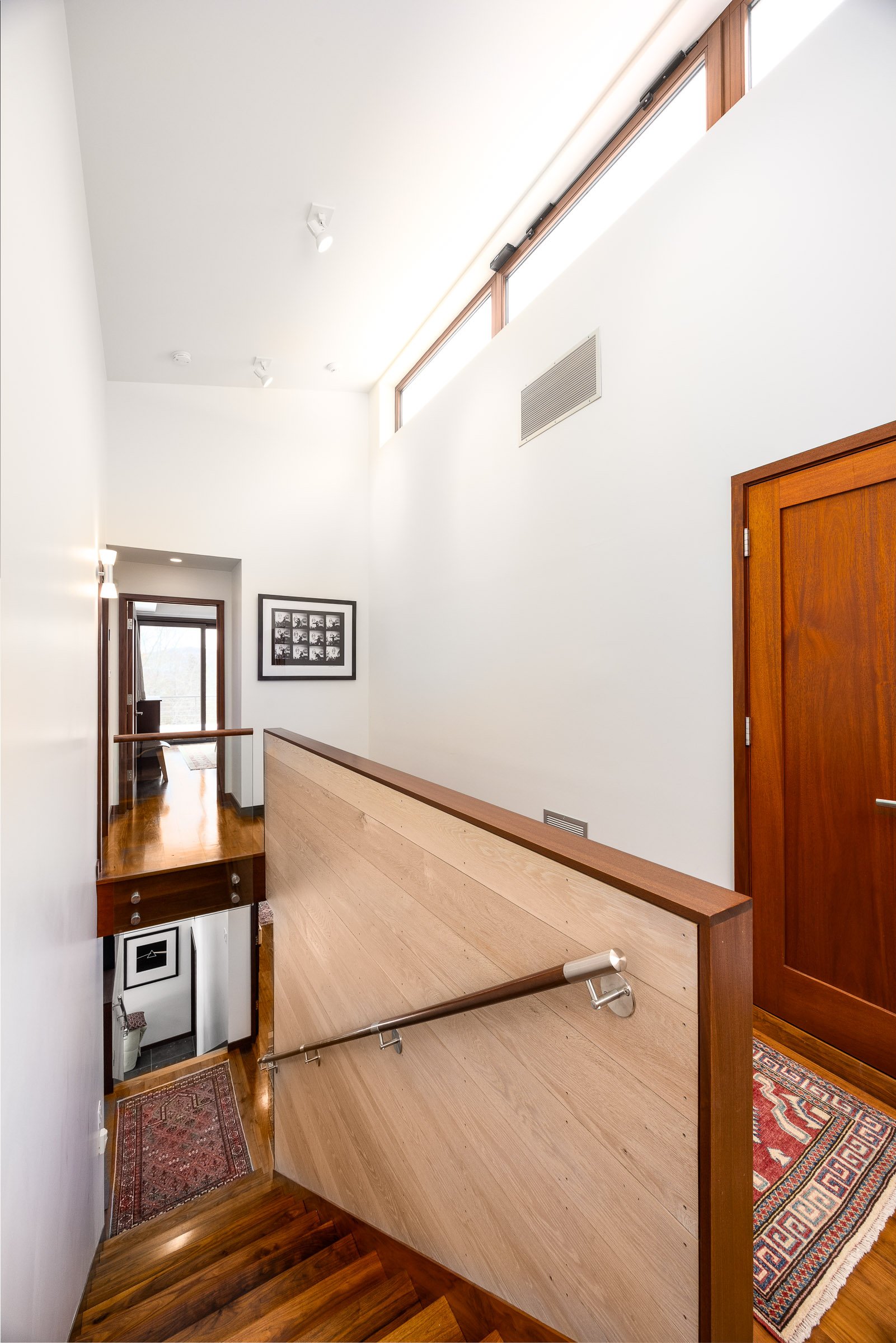Interior view of a modern home staircase landing with wooden floors, white walls, a framed black-and-white photo, high windows, and wooden doors.