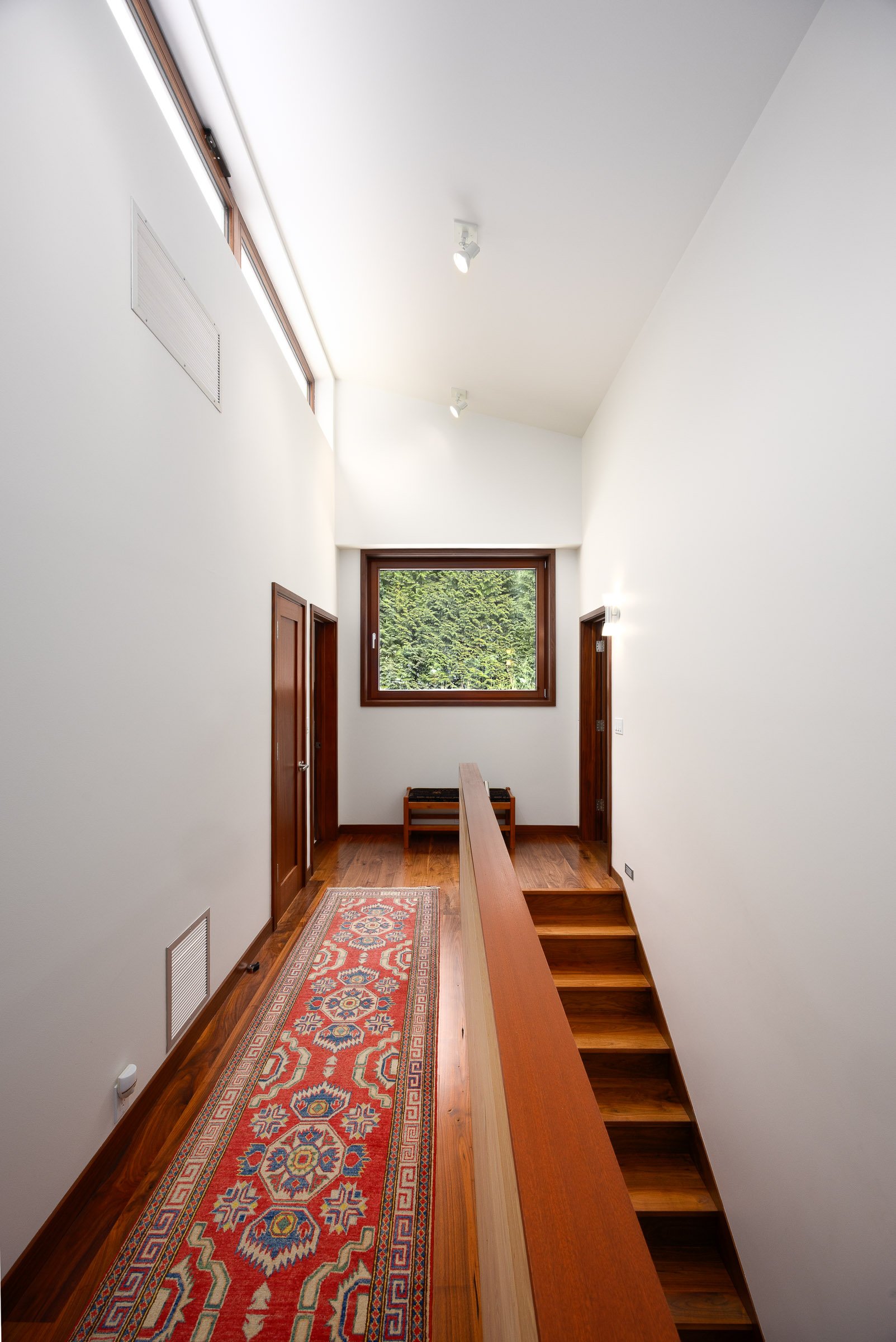 Interior view of a modern staircase area with white walls, wooden floors, small steps leading downward on the right, a window with greenery outside in the background, a patterned runner rug on the floor, and wooden doorways on both sides.