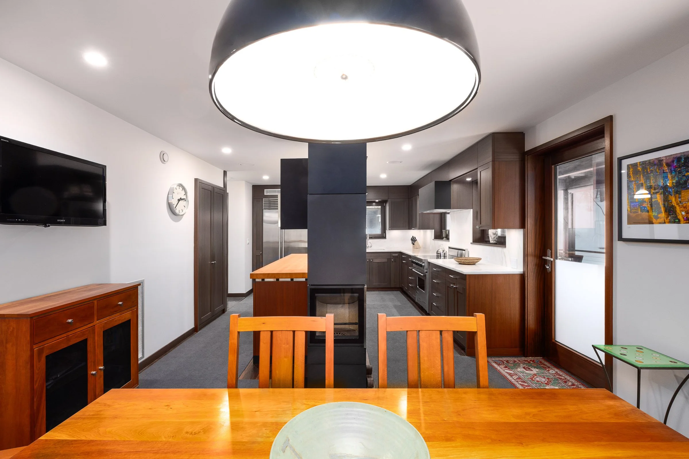 A modern open-plan kitchen and dining area with wooden furniture and dark cabinetry, a large ceiling light, and a wall-mounted TV.