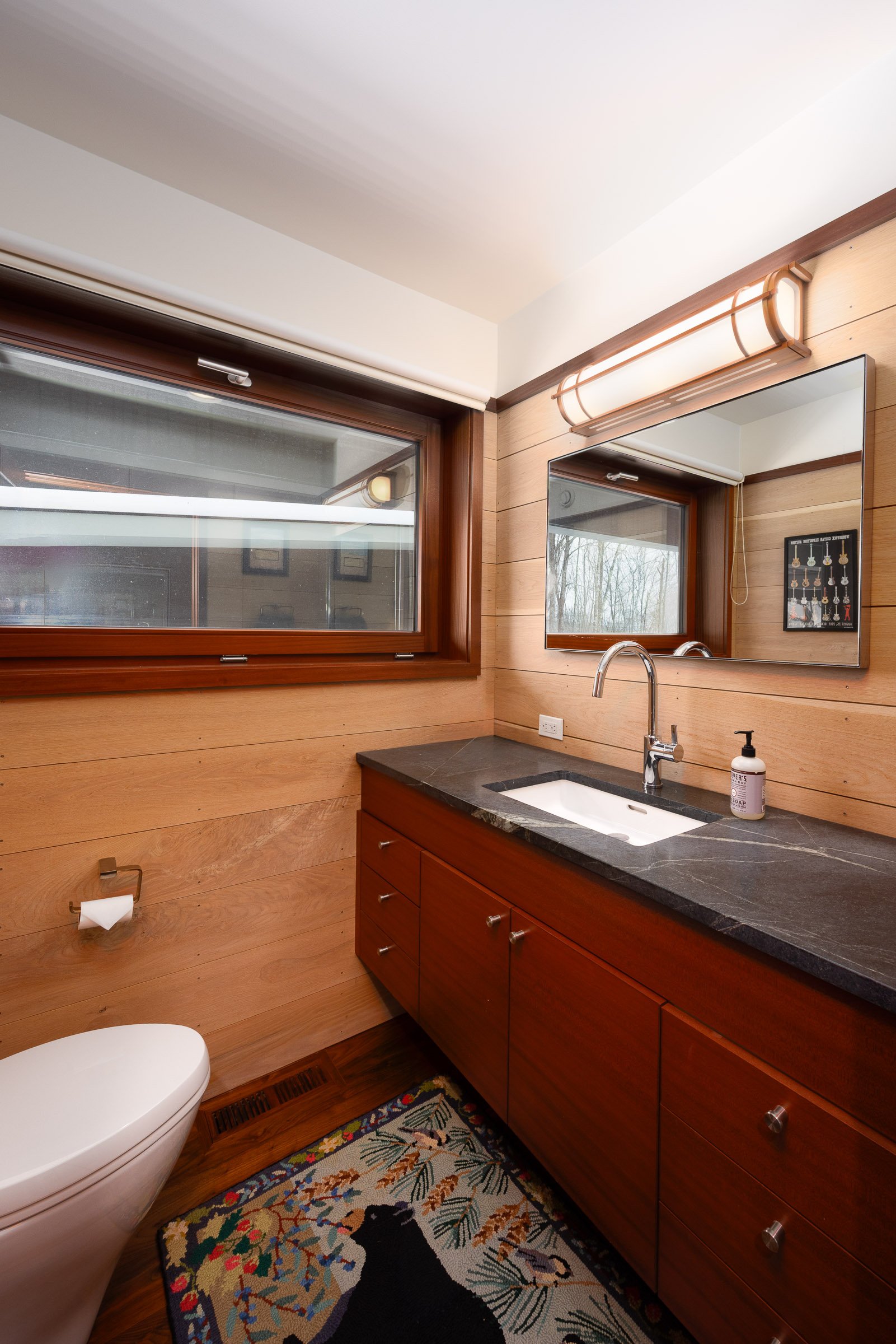 Wood-paneled bathroom with a large mirror, window, black countertop sink, wooden cabinet, decorative rug, and wall-mounted light fixture.