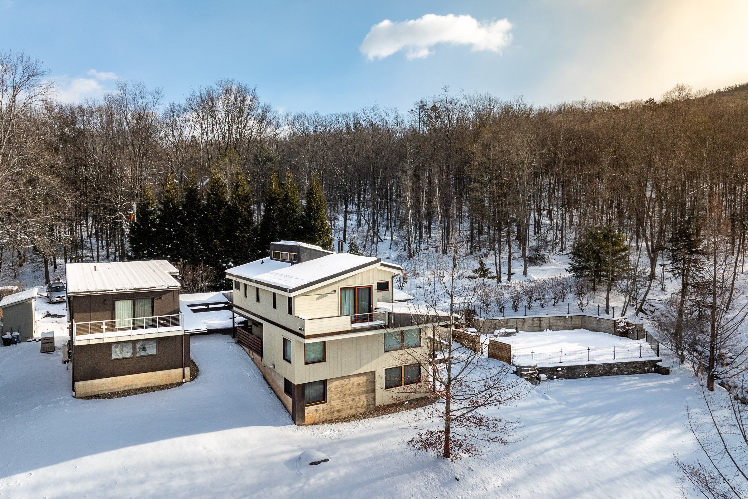 Snow-covered modern house with multiple levels, surrounded by snow and leafless trees, with a wooded hill in the background under a partly cloudy sky.