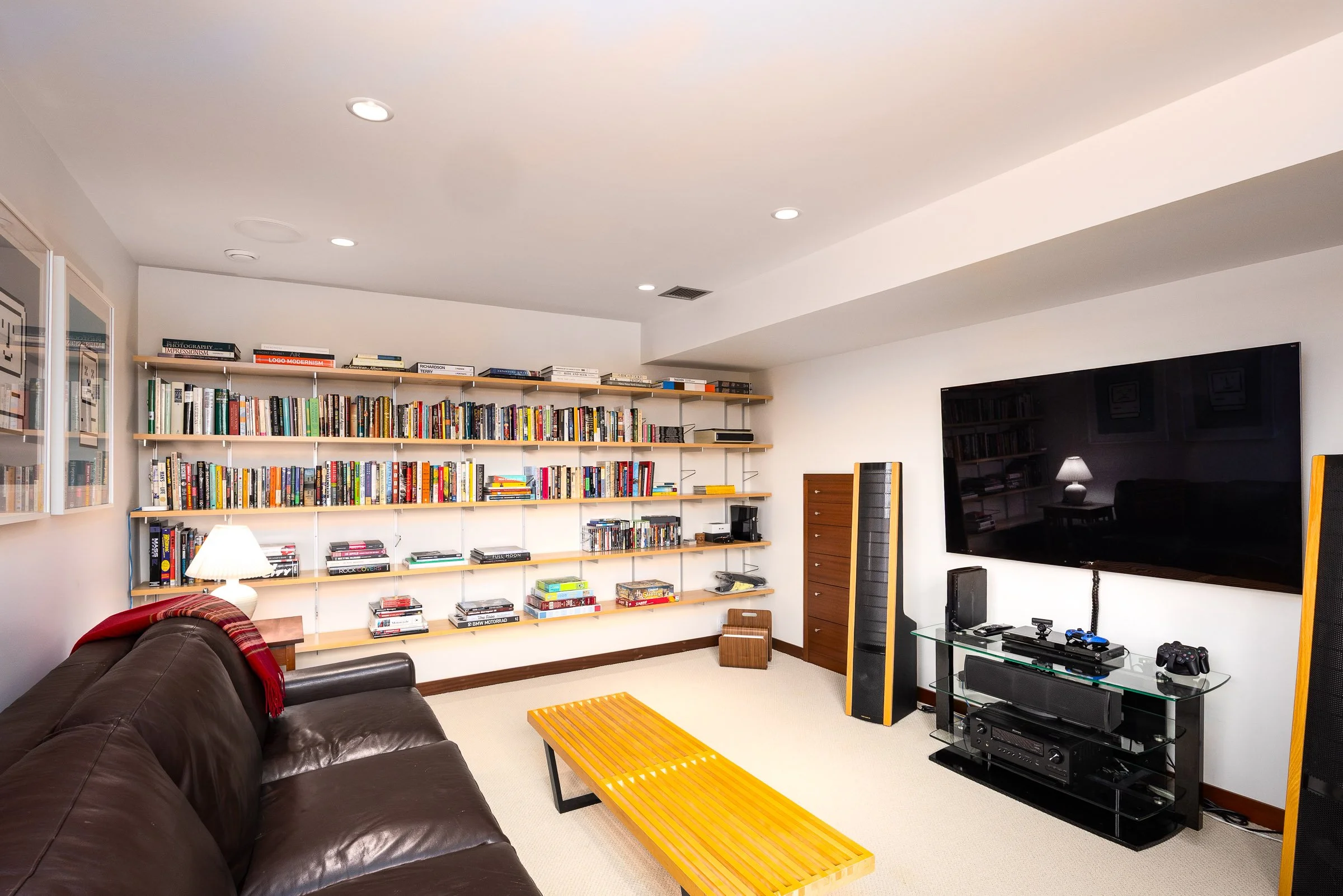Living room with a brown leather sofa, yellow coffee table, white walls, a large flat-screen TV, shelves filled with books, and various electronic devices.