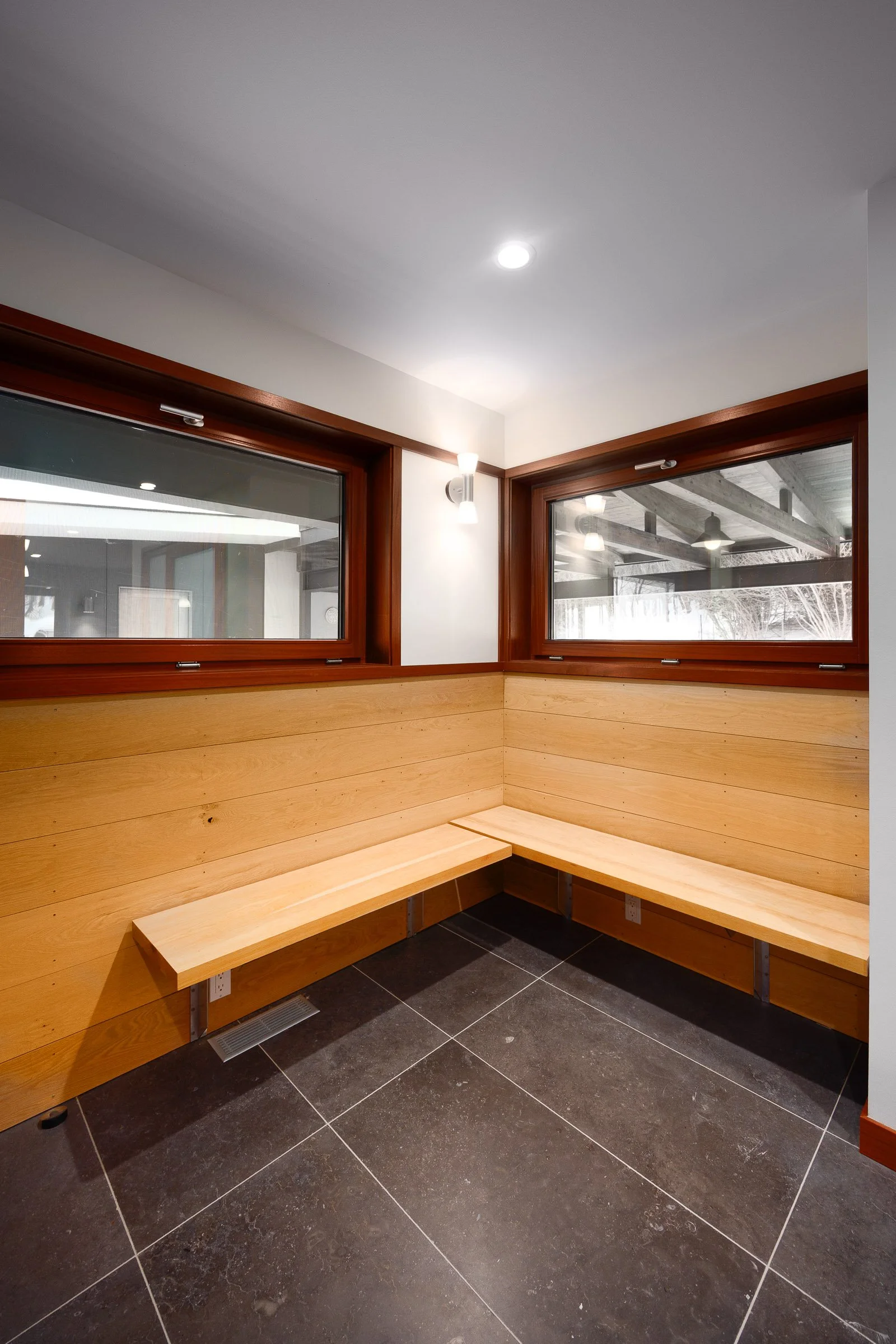 Corner of an indoor room with wooden benches and large windows showing outdoor structures.