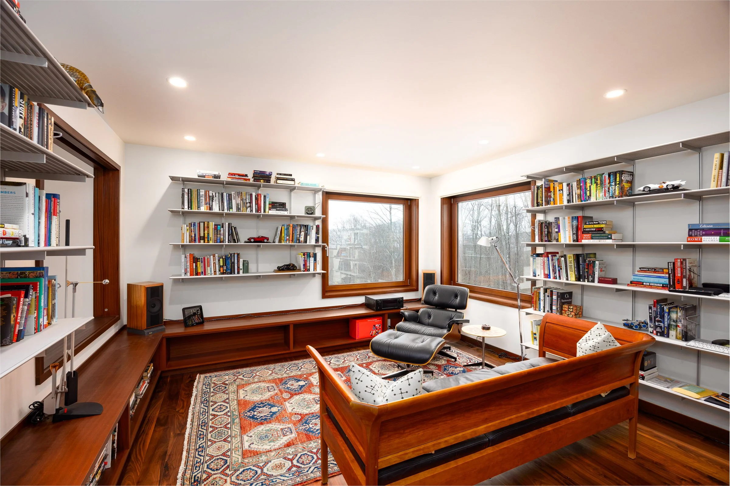 Living room with white walls, large windows, wood trim, and built-in bookshelves filled with books and decorative items. Includes a black lounge chair, a wooden sofa with pillows, a patterned area rug, and various lamps and audio equipment.