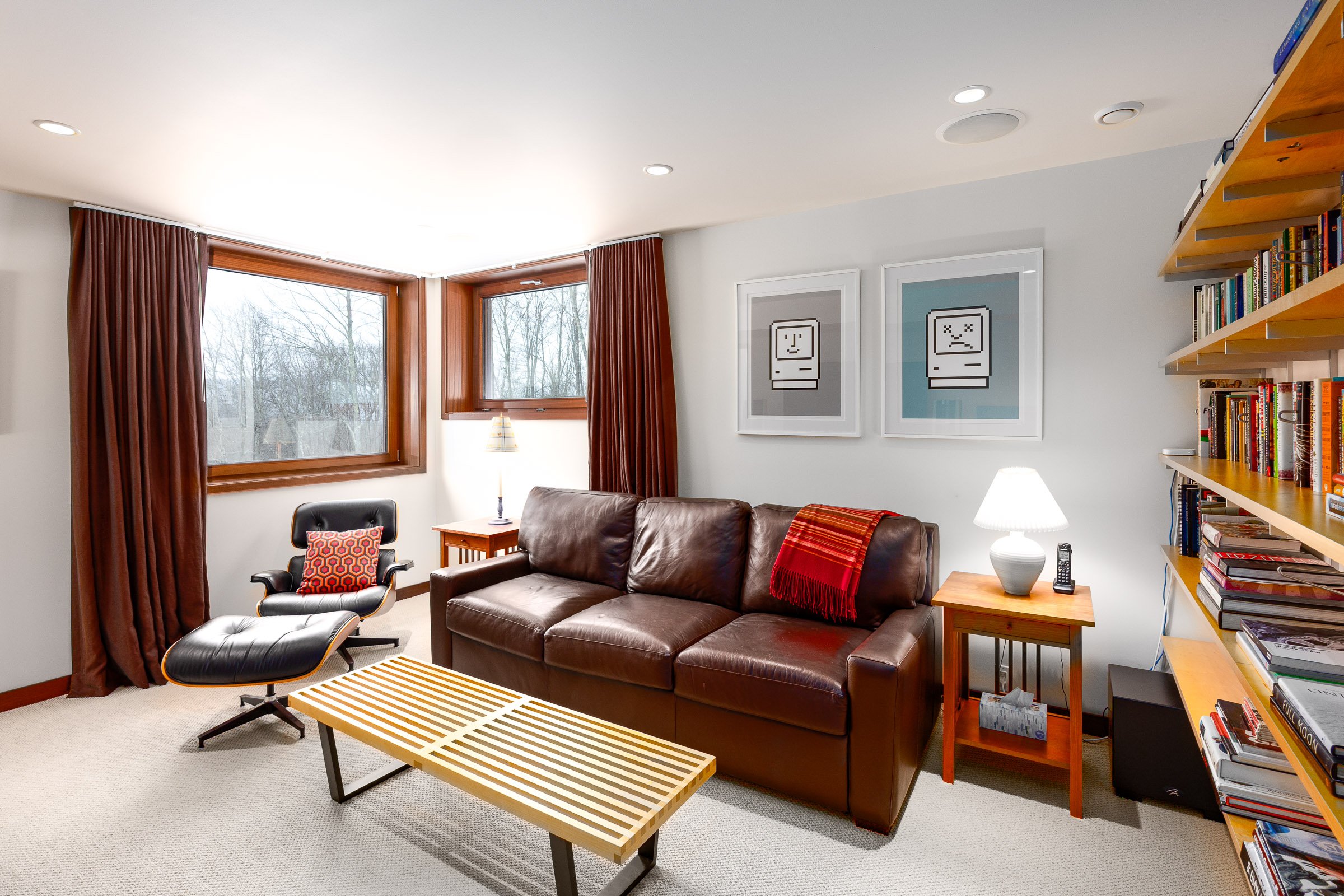 Living room with brown leather sofa, wooden side tables with lamps, black and brown lounge chair, wooden slatted coffee table, wall art with pixelated computer faces, bookshelves filled with books, large windows with wooden trim and brown curtains, b