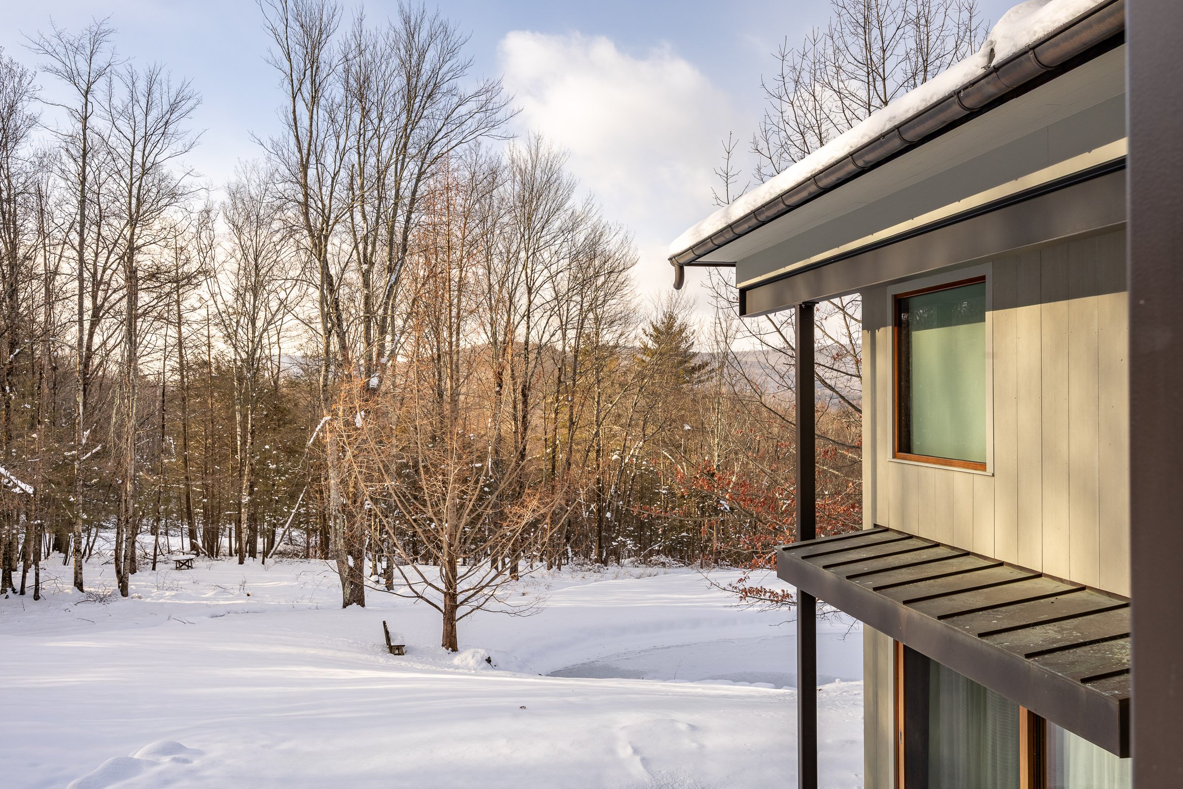 Part of a house with beige siding and wooden window, and a snowy landscape with leafless trees and a bench in the background.