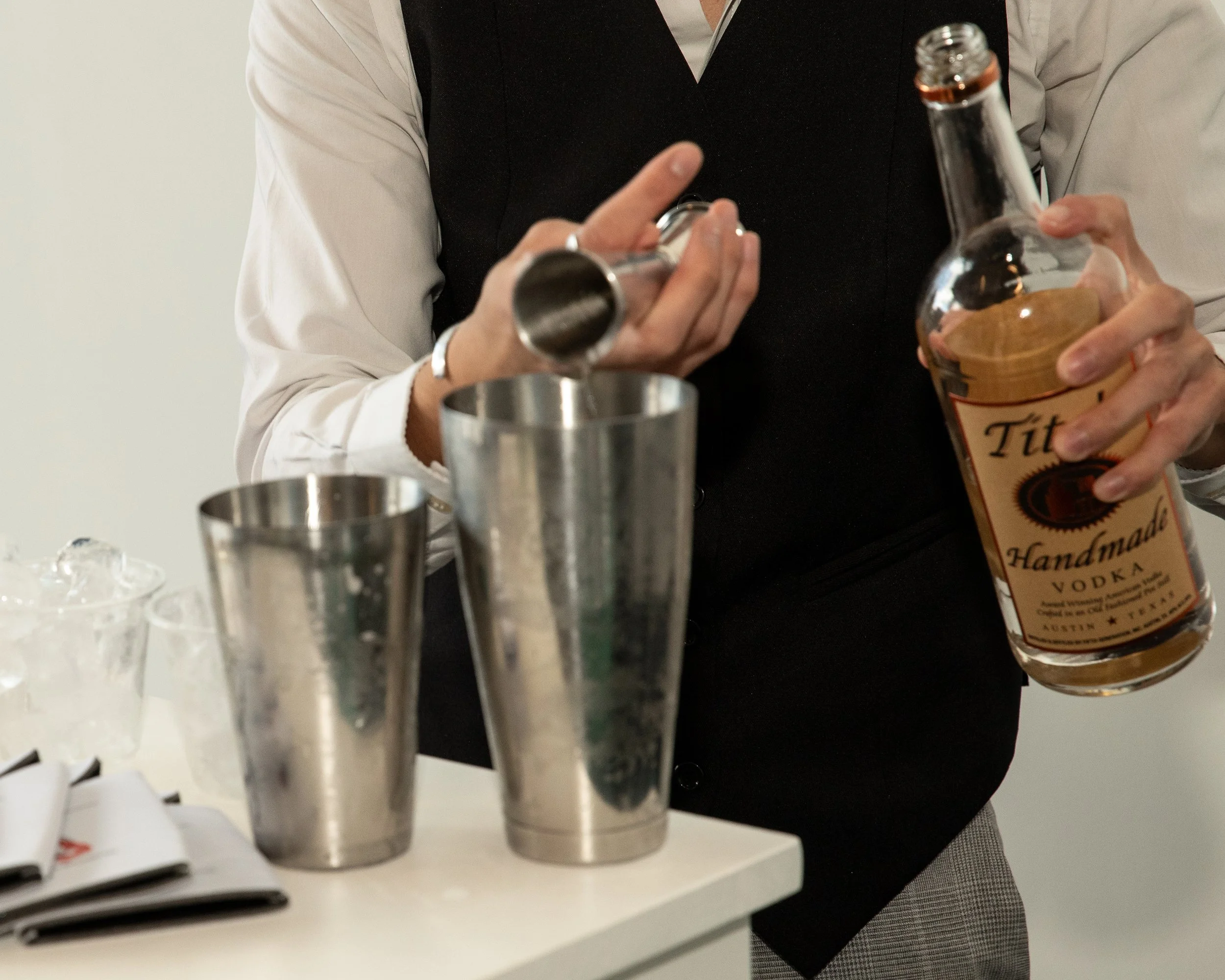 Person pouring vodka into a shaker for making cocktails, with cocktail glasses and bar tools on a white surface.