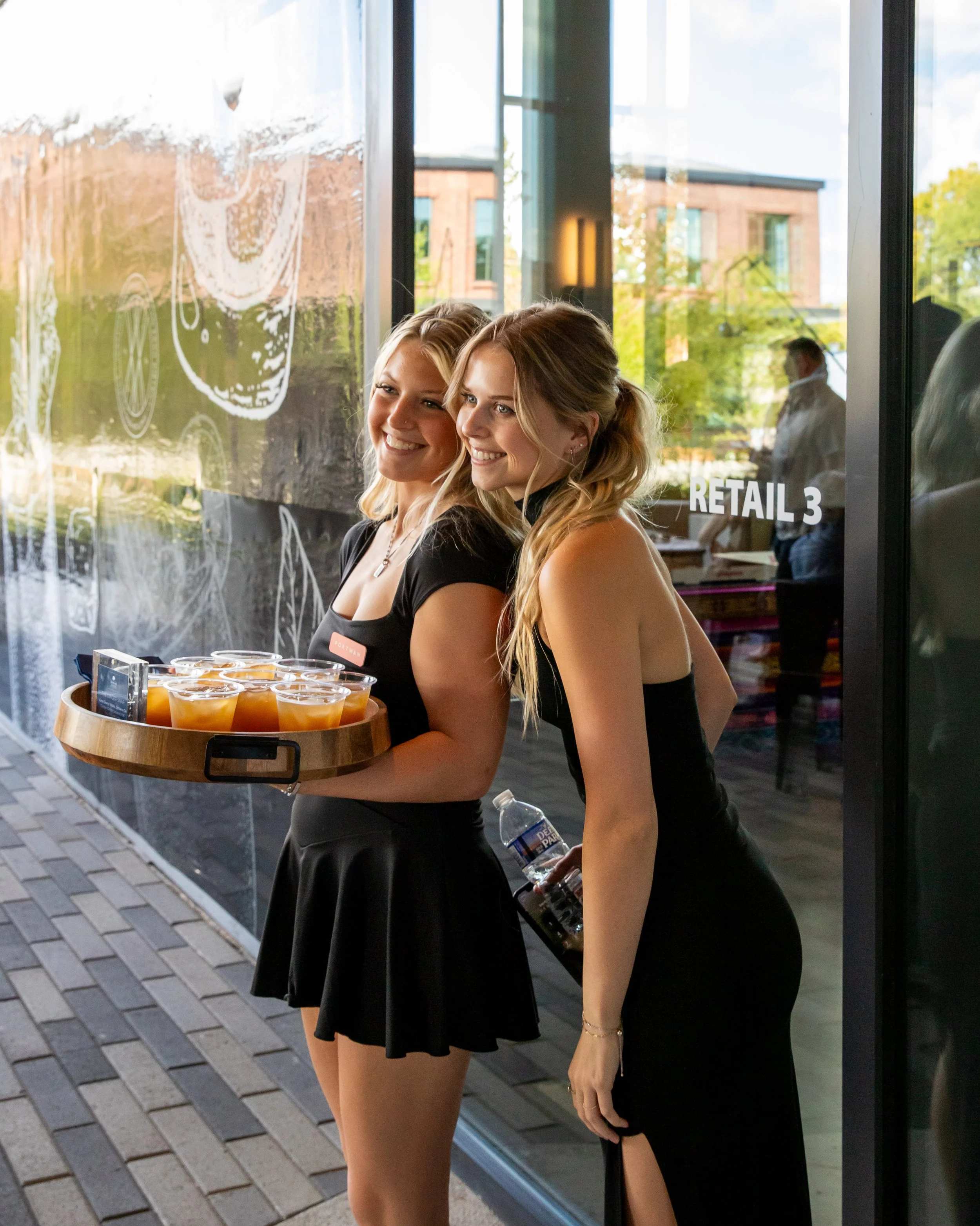 Two smiling young women in black dresses are standing outside a glass storefront. One is holding a tray with cups of orange drinks, and the other is holding a water bottle. They are near a sidewalk with modern buildings in the background.