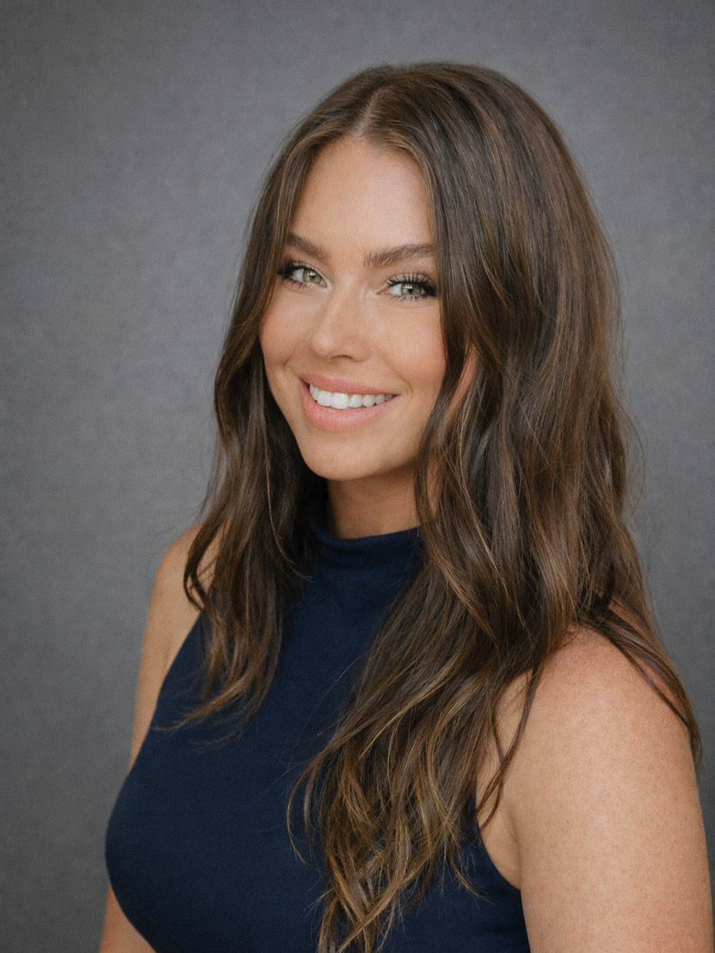 A smiling woman with long, wavy brown hair and light green eyes, wearing a sleeveless navy blue top, standing against a gray background.
