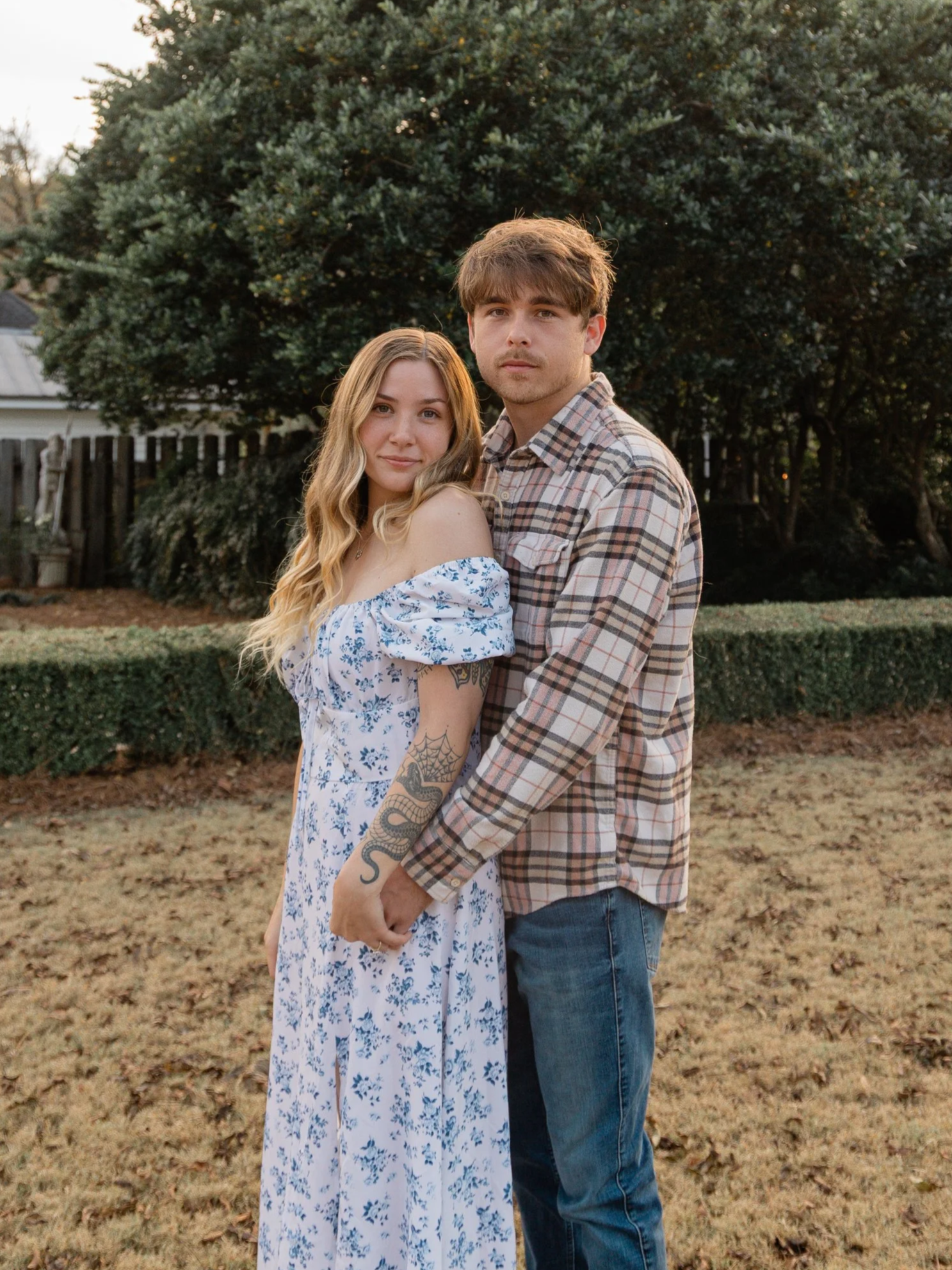 A young couple standing outdoors in a grassy yard, holding hands, with a large tree and a wooden fence in the background during daytime.