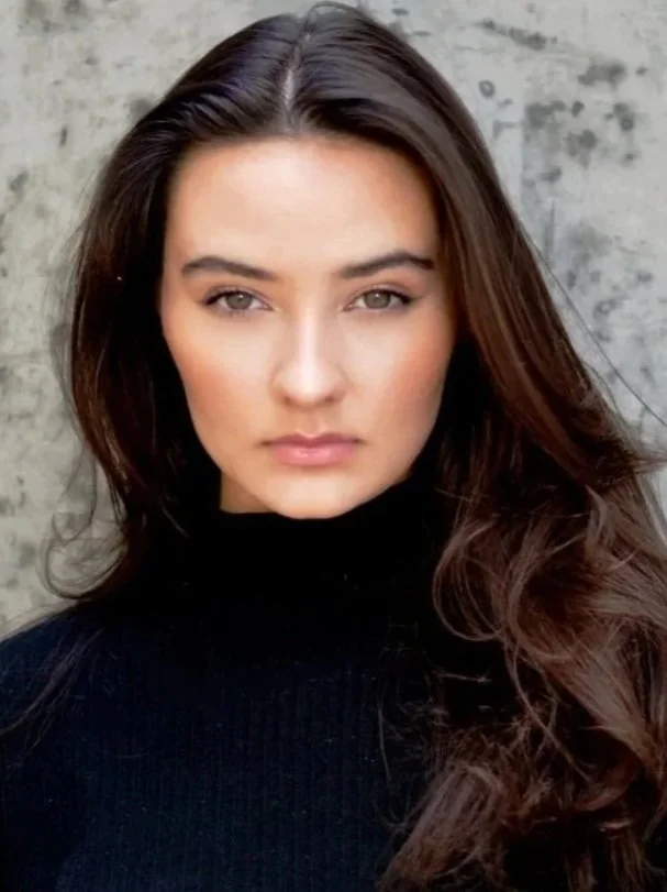 A woman with long, wavy brown hair, wearing a black turtleneck, looking directly at the camera with a serious expression, standing against a gray concrete wall.