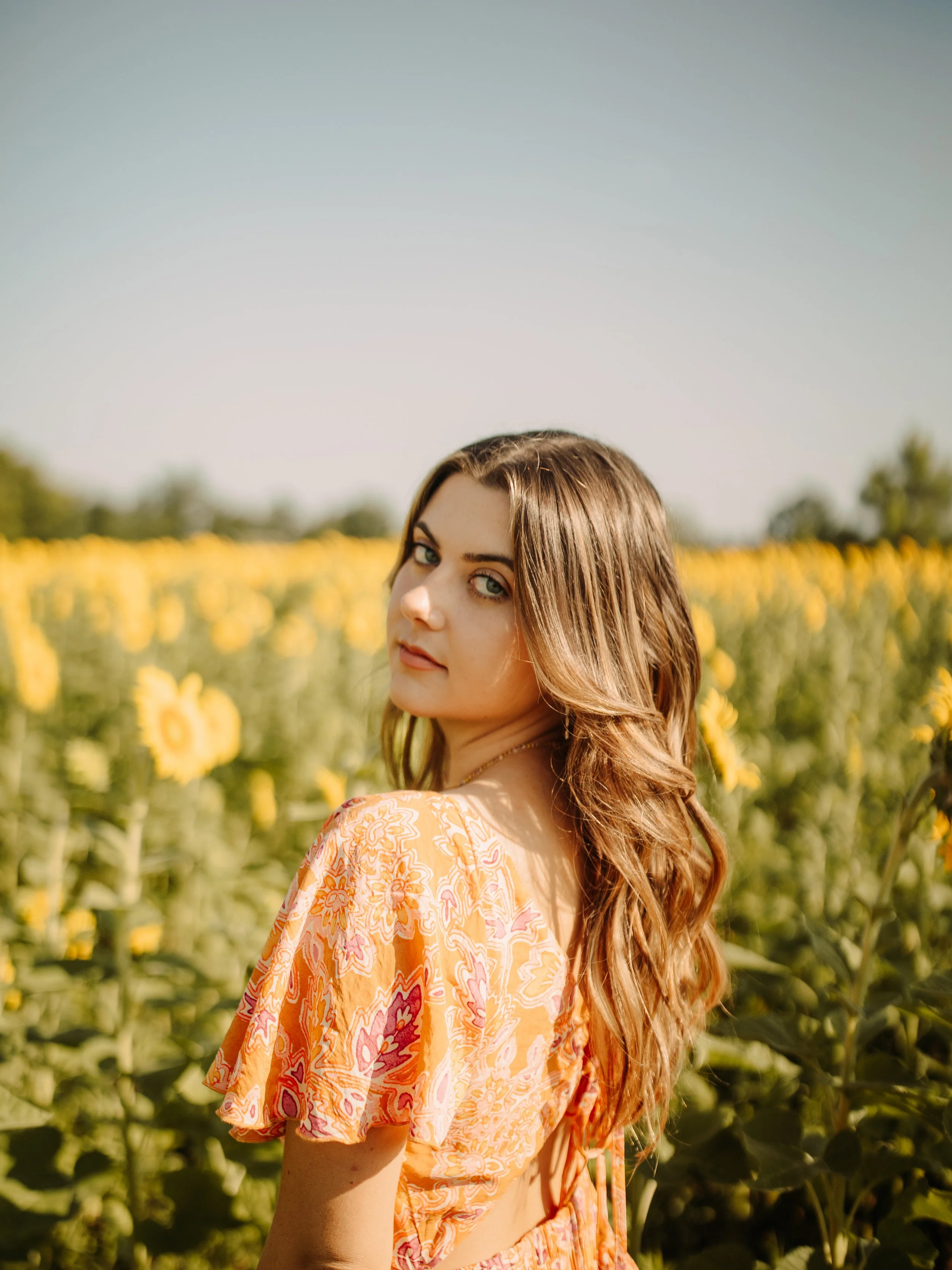 Young woman with long brown hair in a sunflower field wearing an orange patterned dress, looking over her shoulder.