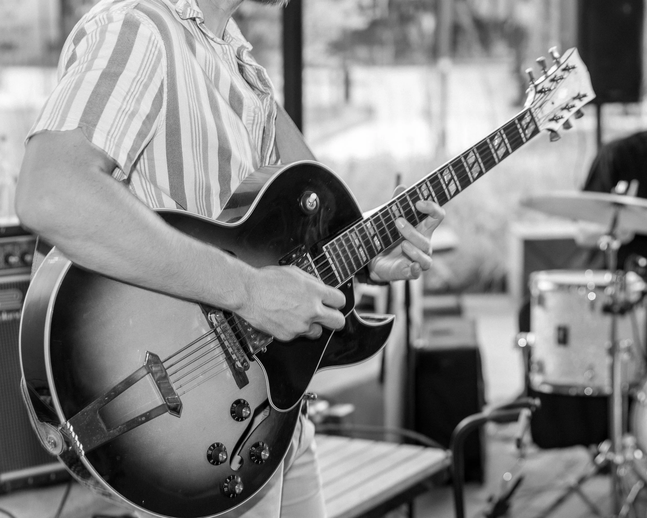 A person playing an electric guitar, wearing a striped shirt, in a room with musical equipment in the background.