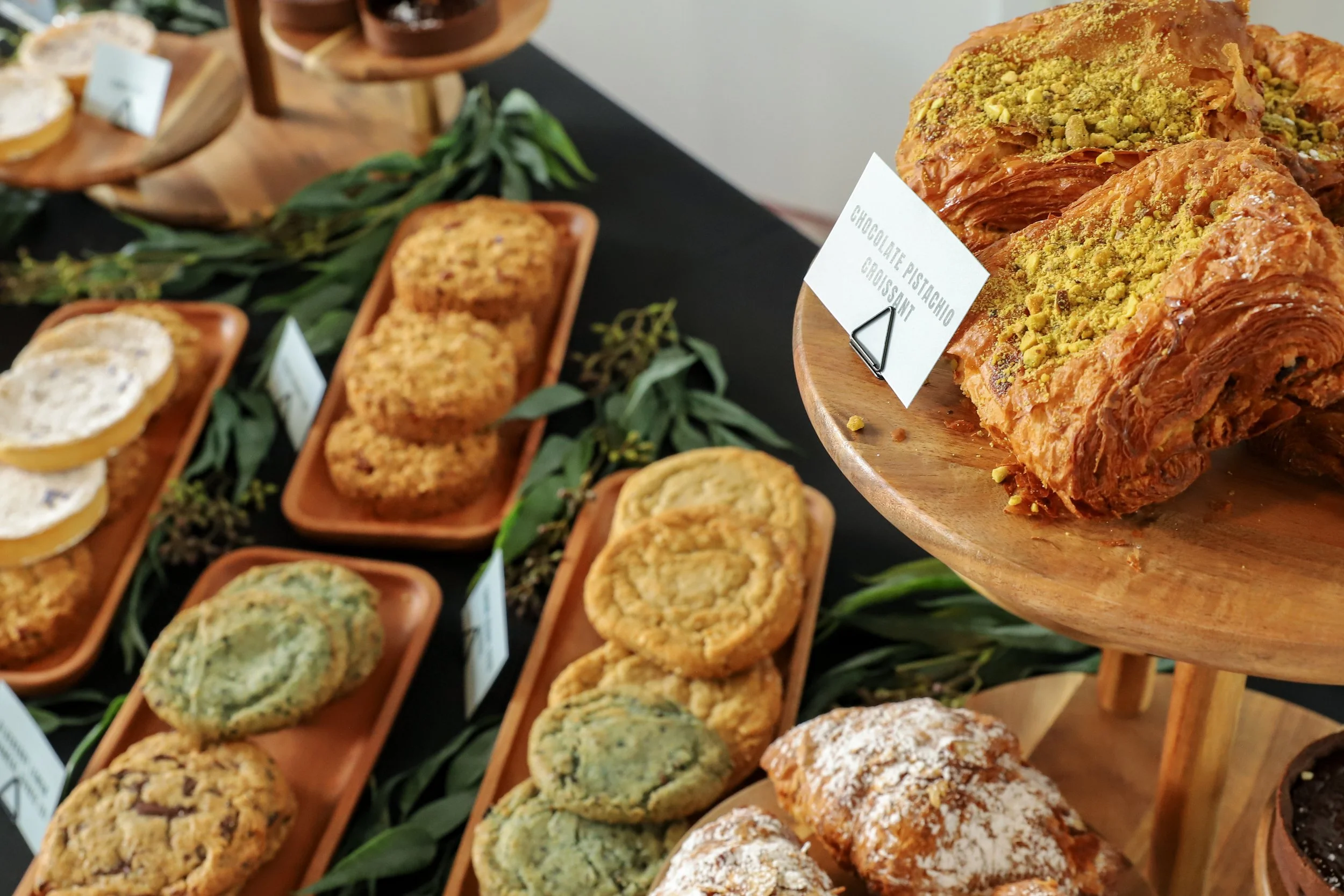 Assorted baked goods including croissants topped with pistachios, cookies, and pastries displayed on wooden trays and stands with small labels.