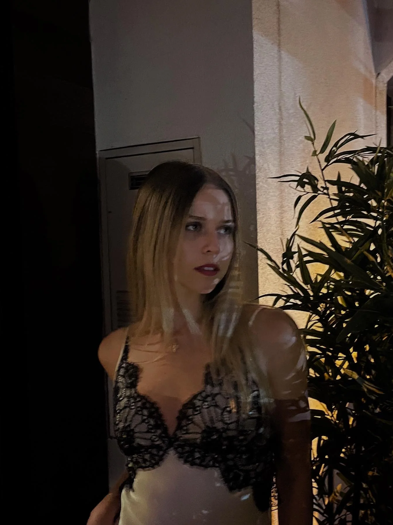 A woman with blonde hair wearing a black lace top standing near a wall and a plant at night.