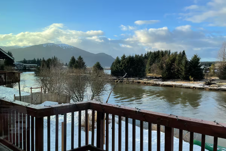 View from a wooden deck overlooking a river with snow on the banks, a small island with trees, mountains in the background, and a partly cloudy sky.