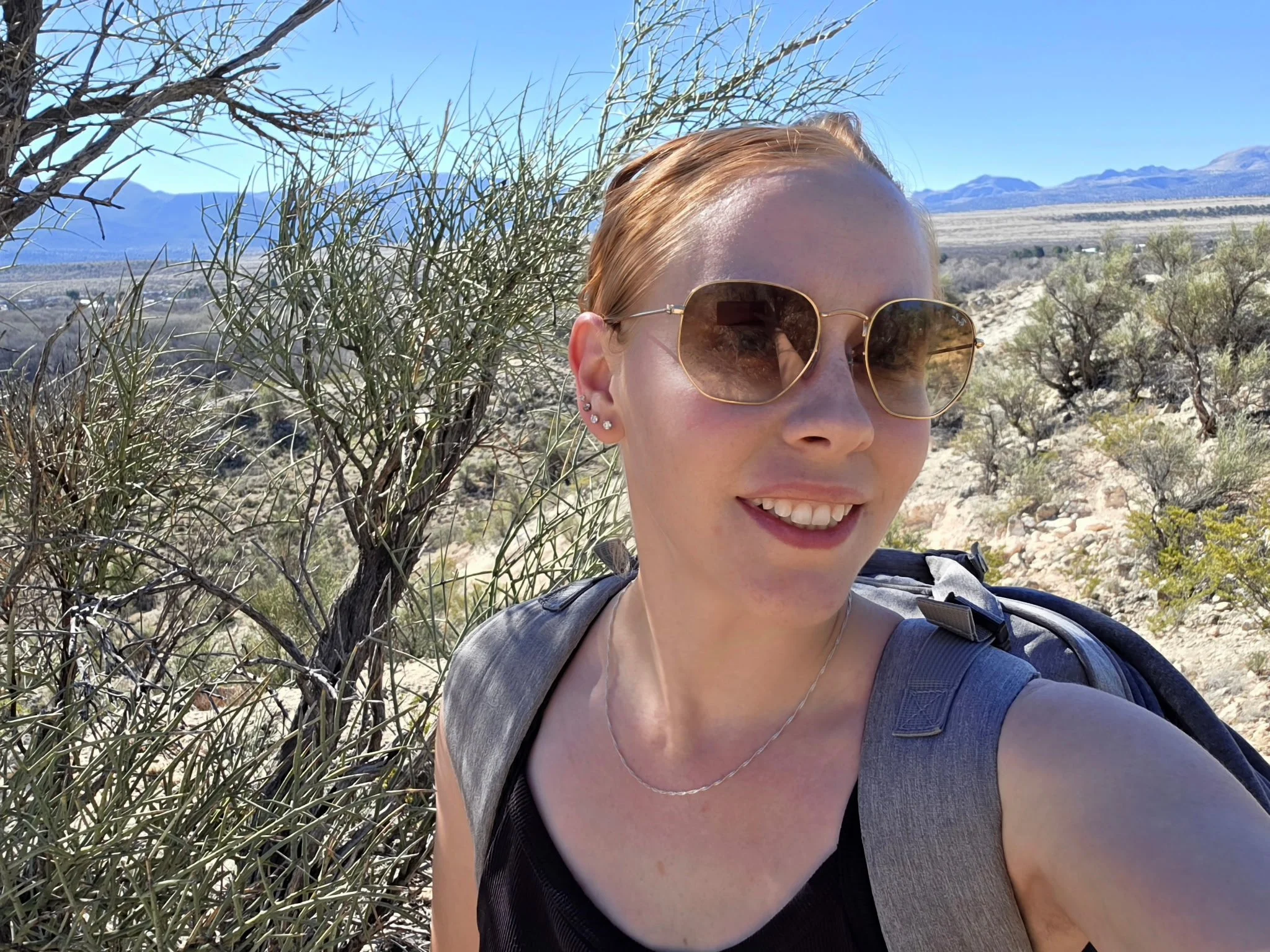 A woman with blonde hair wearing sunglasses and a black top, smiling outdoors in a desert landscape with shrubs, mountains, and a clear blue sky.