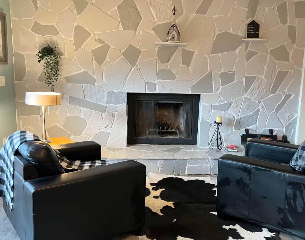 Living room with a stone fireplace, two black leather armchairs, a white lamp, a black and white checkered blanket, a black cowhide rug, a small side table with a candle, and decorative shelves with small ornaments on a textured stone wall.
