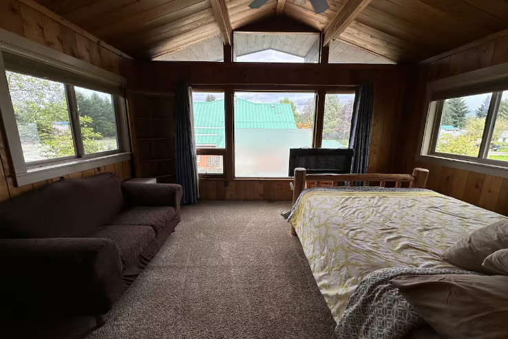 A cozy bedroom with wood-paneled walls and ceiling, a sofa on the left, a bed on the right, and large windows with a view of trees and rooftops outside.
