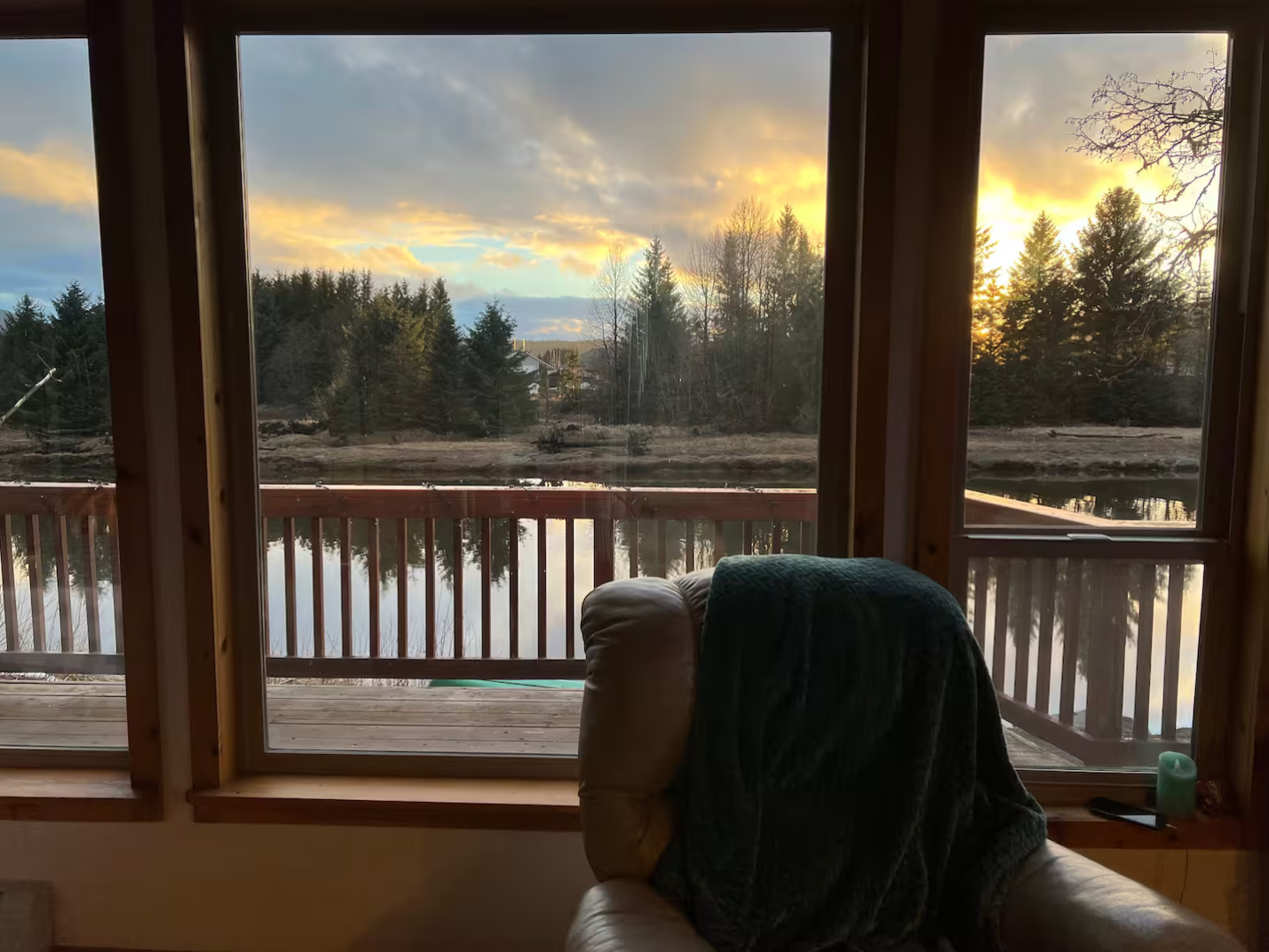 Sunset view of trees and water through a wooden framed window, with a cozy chair in the foreground.