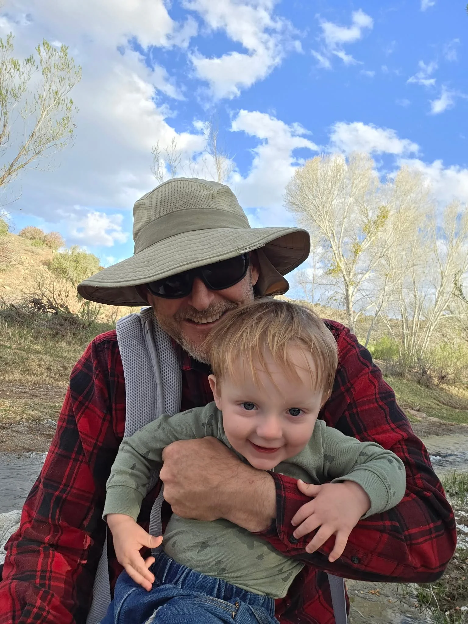 A man wearing a wide-brimmed hat and sunglasses holding a smiling young boy outdoors with trees and blue sky in the background.