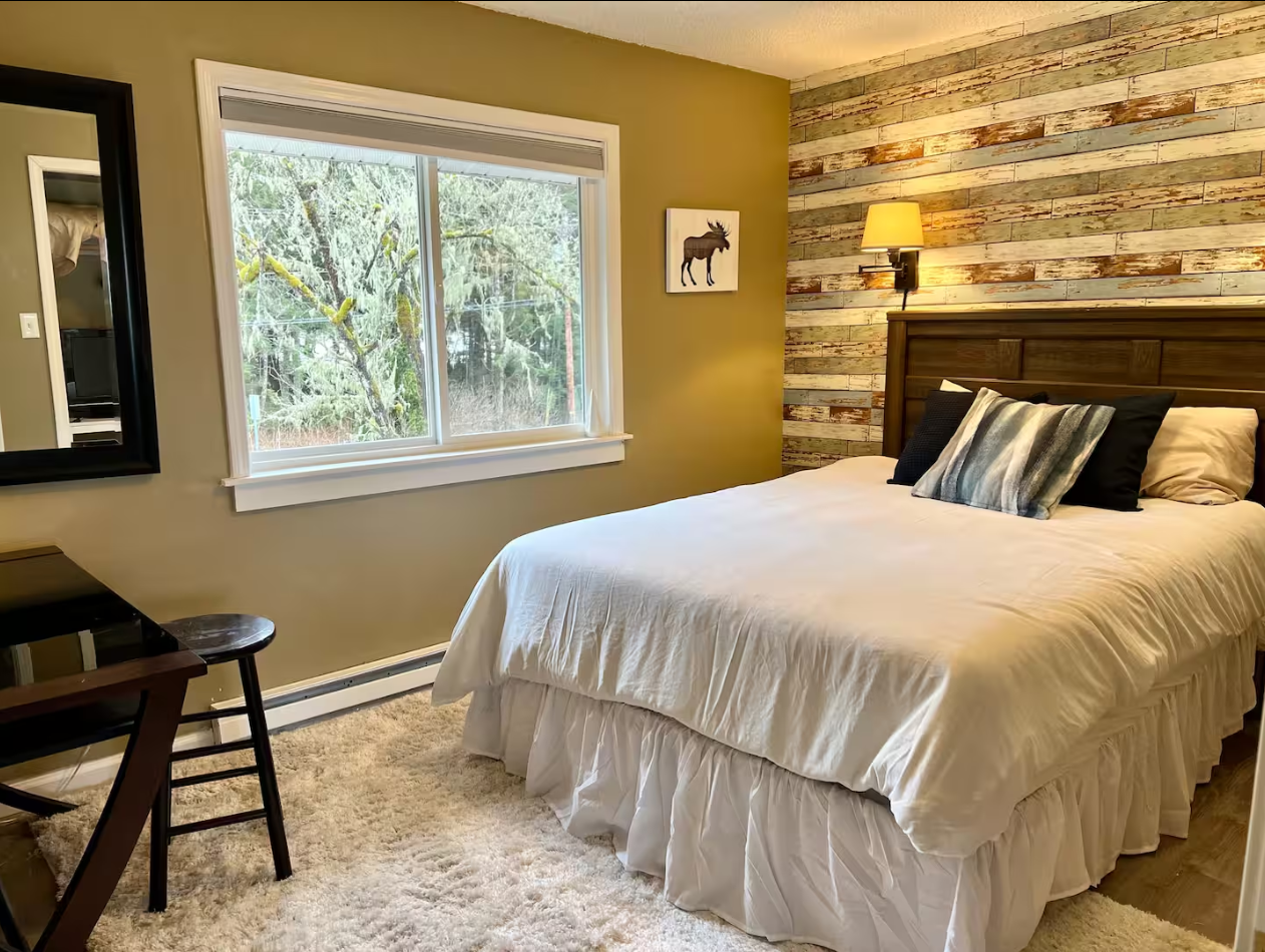 A cozy bedroom with a bed featuring white bedding and decorative pillows, a rustic wooden headboard, a wall-mounted lamp, a window with a view of trees, and a wall with wooden planks. There is also a mirror, a small desk, and a chair.