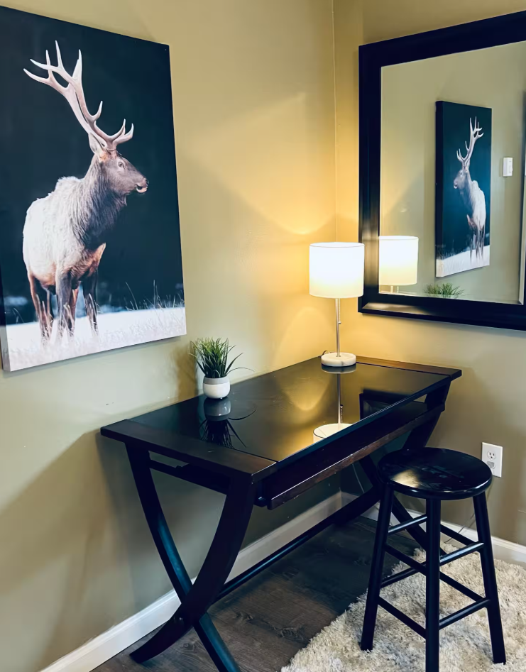 Decoration corner with a black desk, a tall black-stool, a lamp, small plant, and wall art of a deer with large antlers reflected in a mirror.