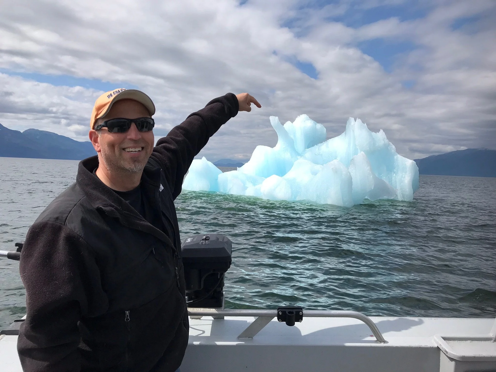 Man smiling on boat pointing at iceberg in water, with mountains and cloudy sky in background.