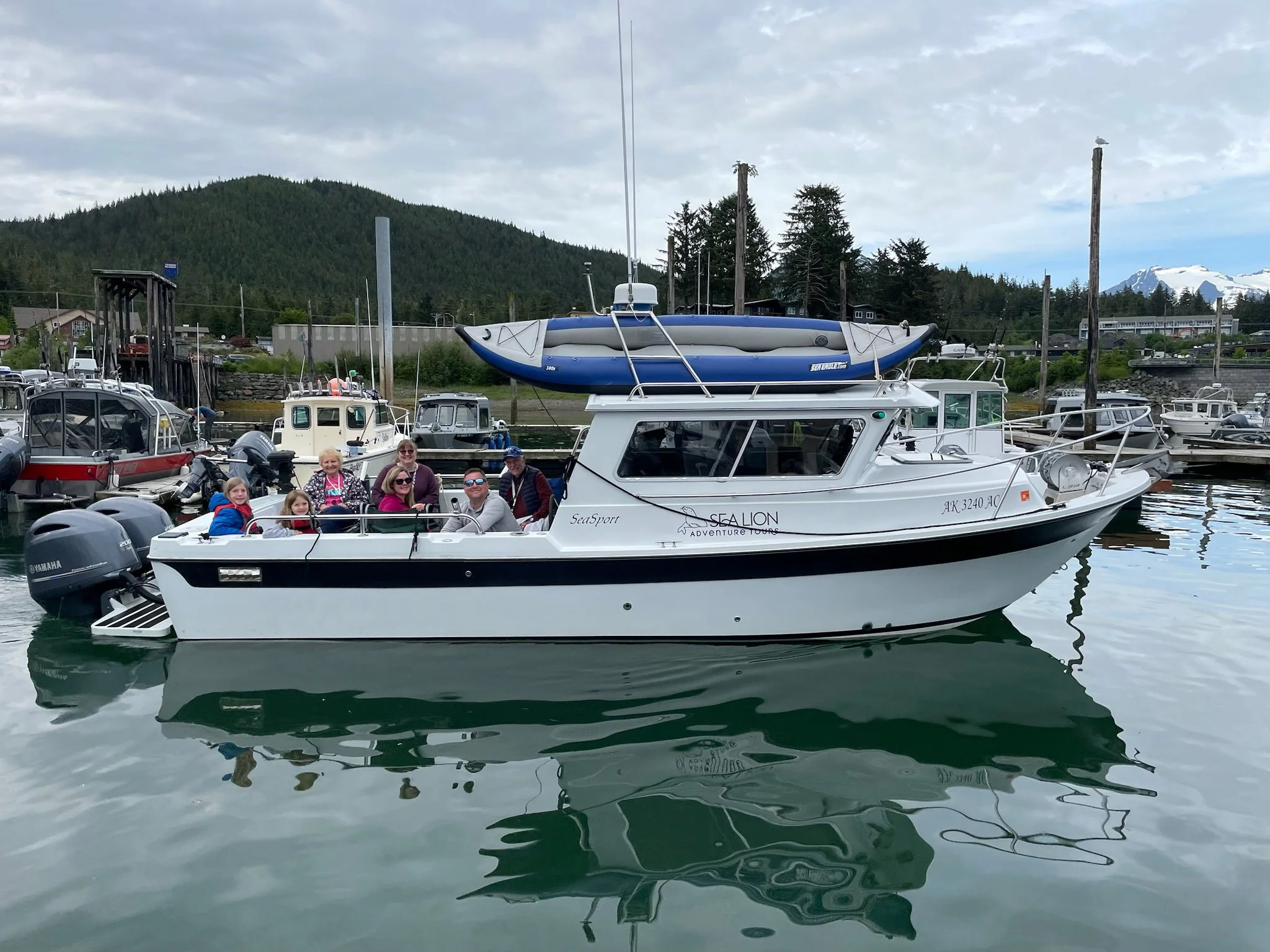 Group of people on a white and black boat named SEALION docked at a marina with other boats, trees, hills, and mountains in the background.