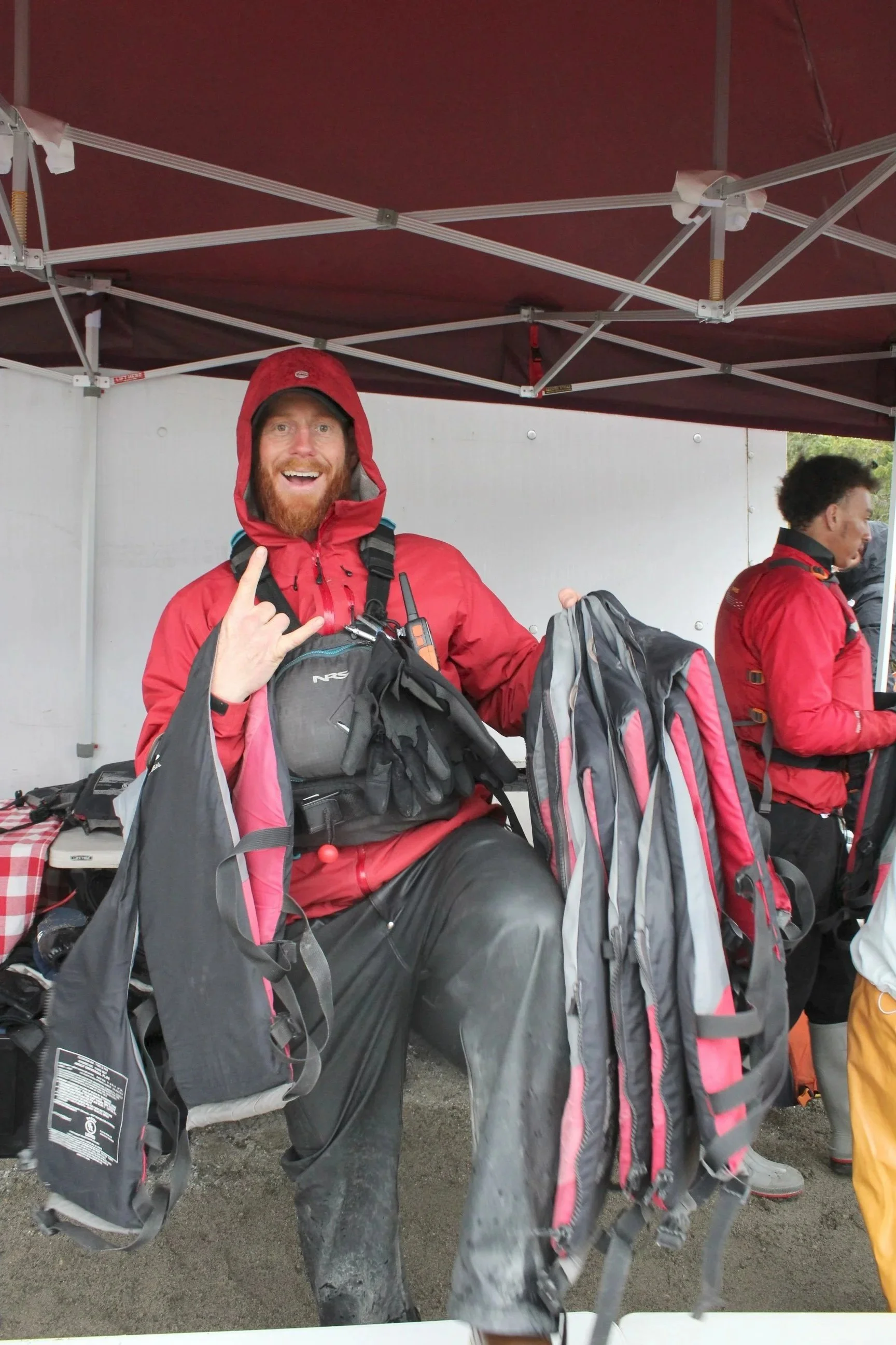 Man in red rain jacket and hood smiling and making a rock on hand gesture while sitting under a maroon canopy, surrounded by backpacks and gear.
