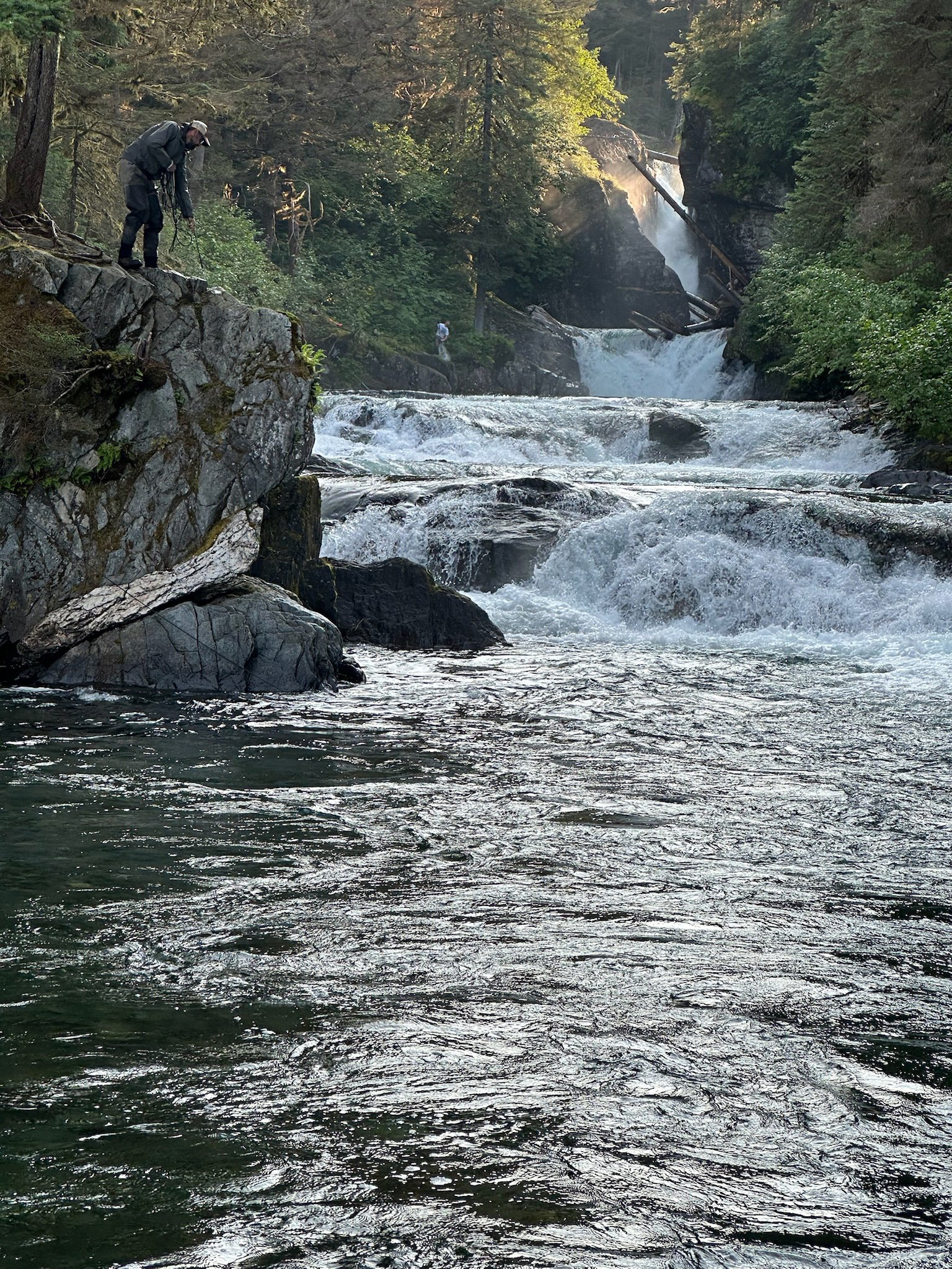 A person in outdoor gear is preparing to climb a large rock beside a flowing river with small waterfalls, surrounded by green trees and cliffs.