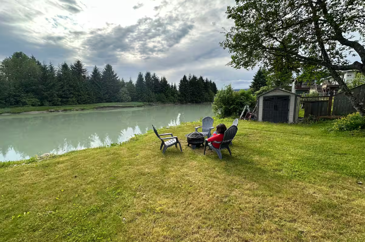 A backyard with four black chairs around a fire pit, a person sitting in one chair, facing a body of water, with trees and cloudy sky in the background.