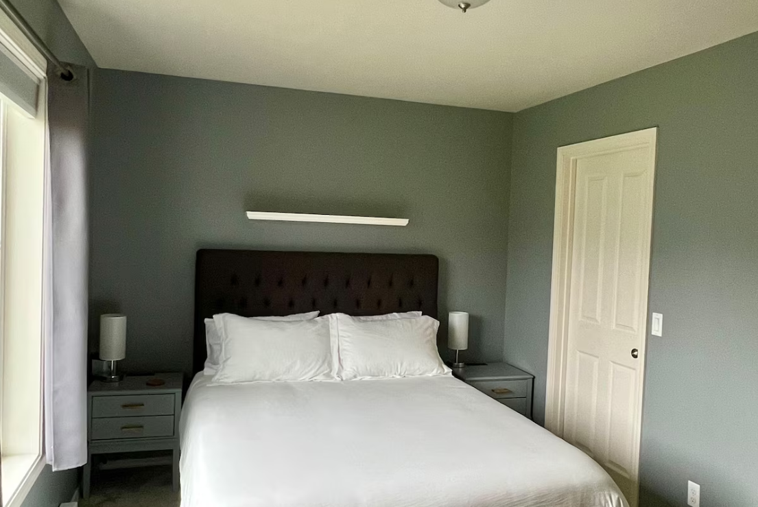 A bedroom with a bed, gray walls, two nightstands, lamps, a door, and a window with curtains.