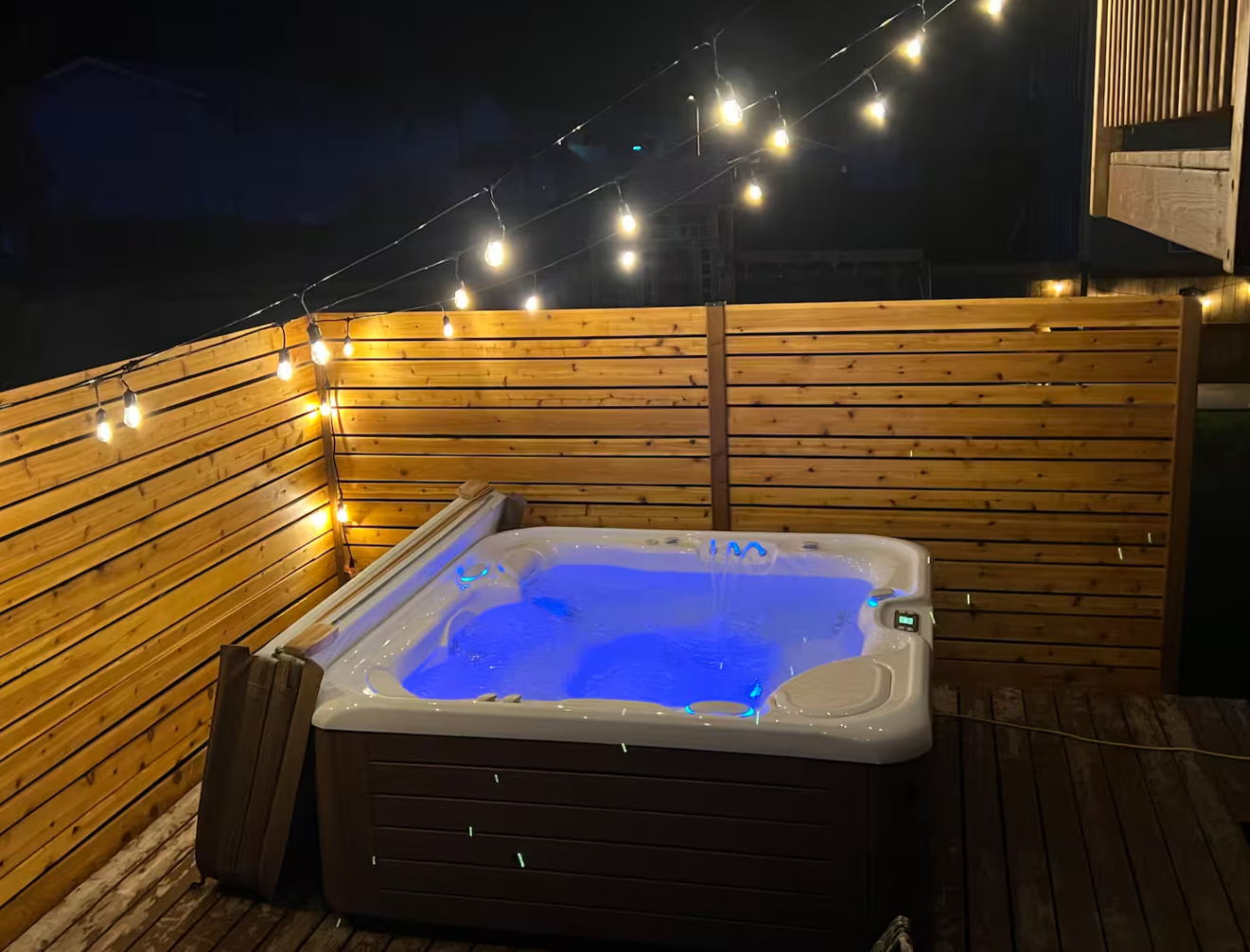 An outdoor hot tub with blue-lit water on a wooden deck, surrounded by a wooden fence, illuminated by string lights at night.