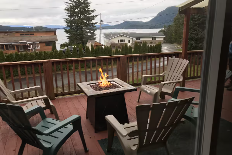 Backyard deck with four Adirondack chairs around a small fire pit, overlooking a neighborhood with houses, trees, a body of water, and mountains in the background on a cloudy day.