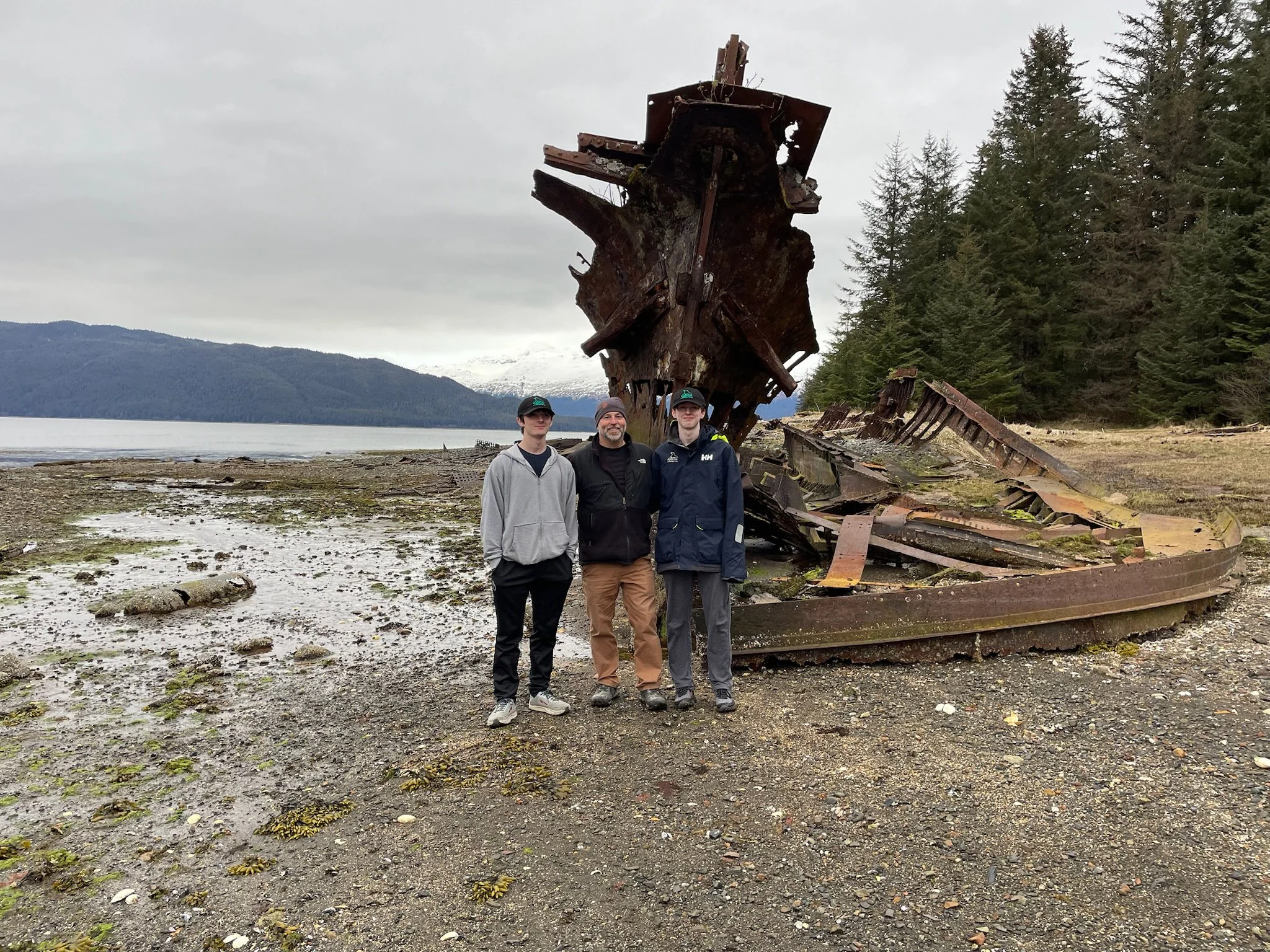 Three people stand in front of a large rusty, wrecked ship on a beach with water, mountains, and trees in the background.