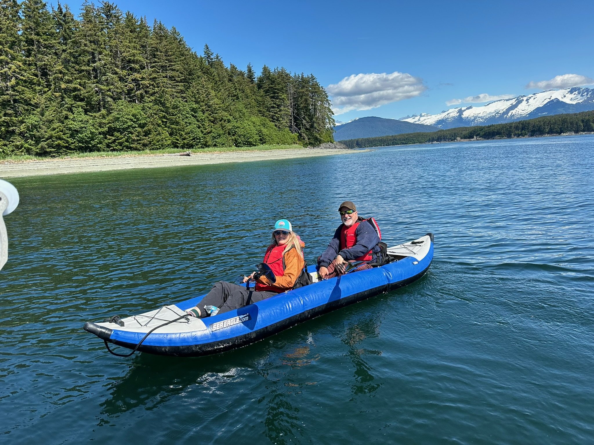 Two people in a blue inflatable kayak on a lake with trees on the shore and snow-capped mountains in the background, under a partly cloudy sky.