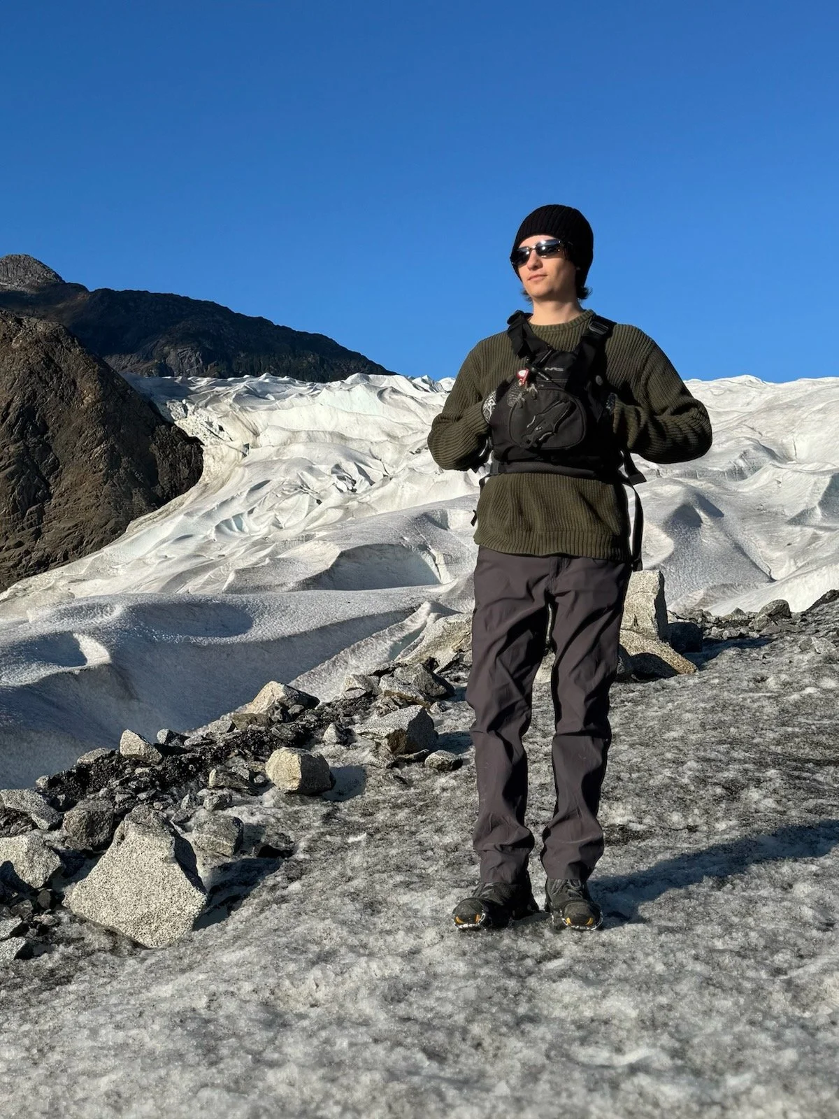 A person dressed in outdoor winter gear, standing on snow and ice with a glacier and mountain in the background under a clear blue sky.